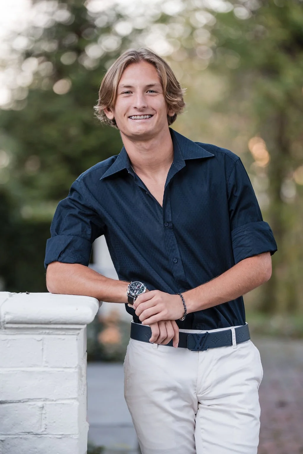 Young man with blonde hair smiling, standing outdoors, wearing a navy blue shirt and white pants, leaning on a white stone railing.