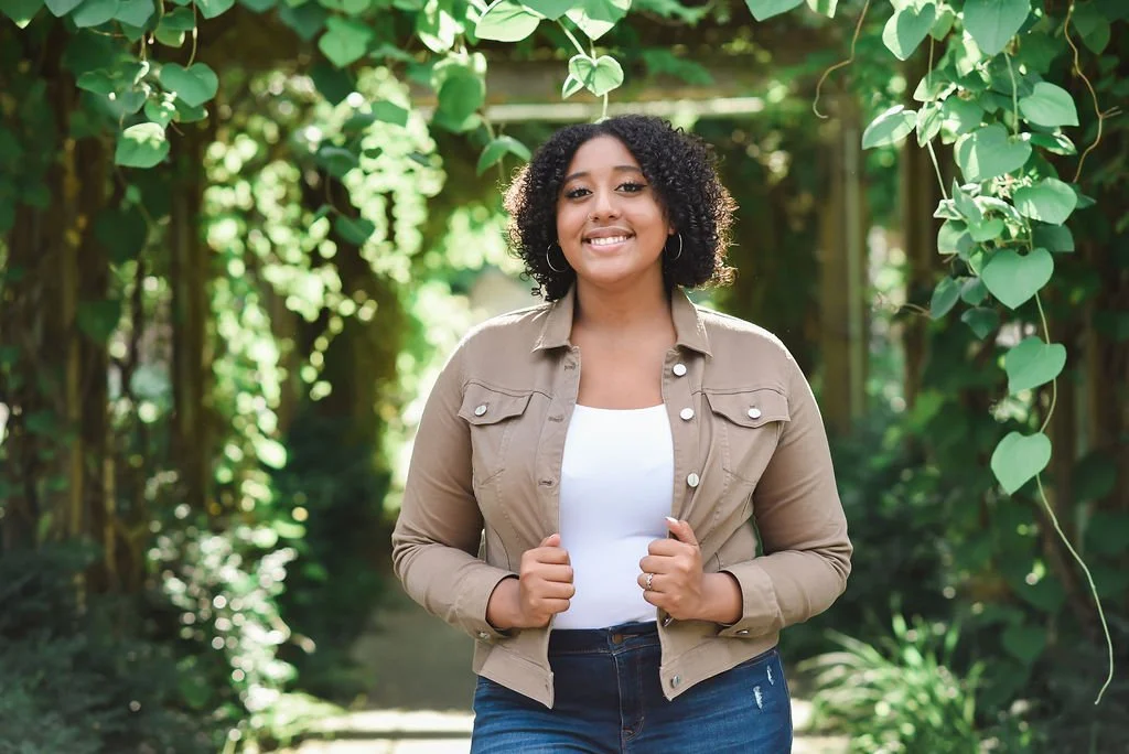 A young woman with curly hair smiling and standing outdoors in a lush, green garden or park.