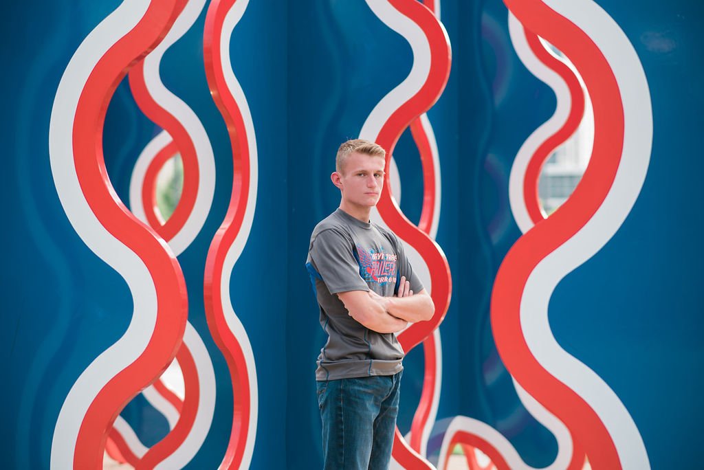 Young man standing with crossed arms in front of a colorful abstract wall with wavy red, white, and blue patterns.