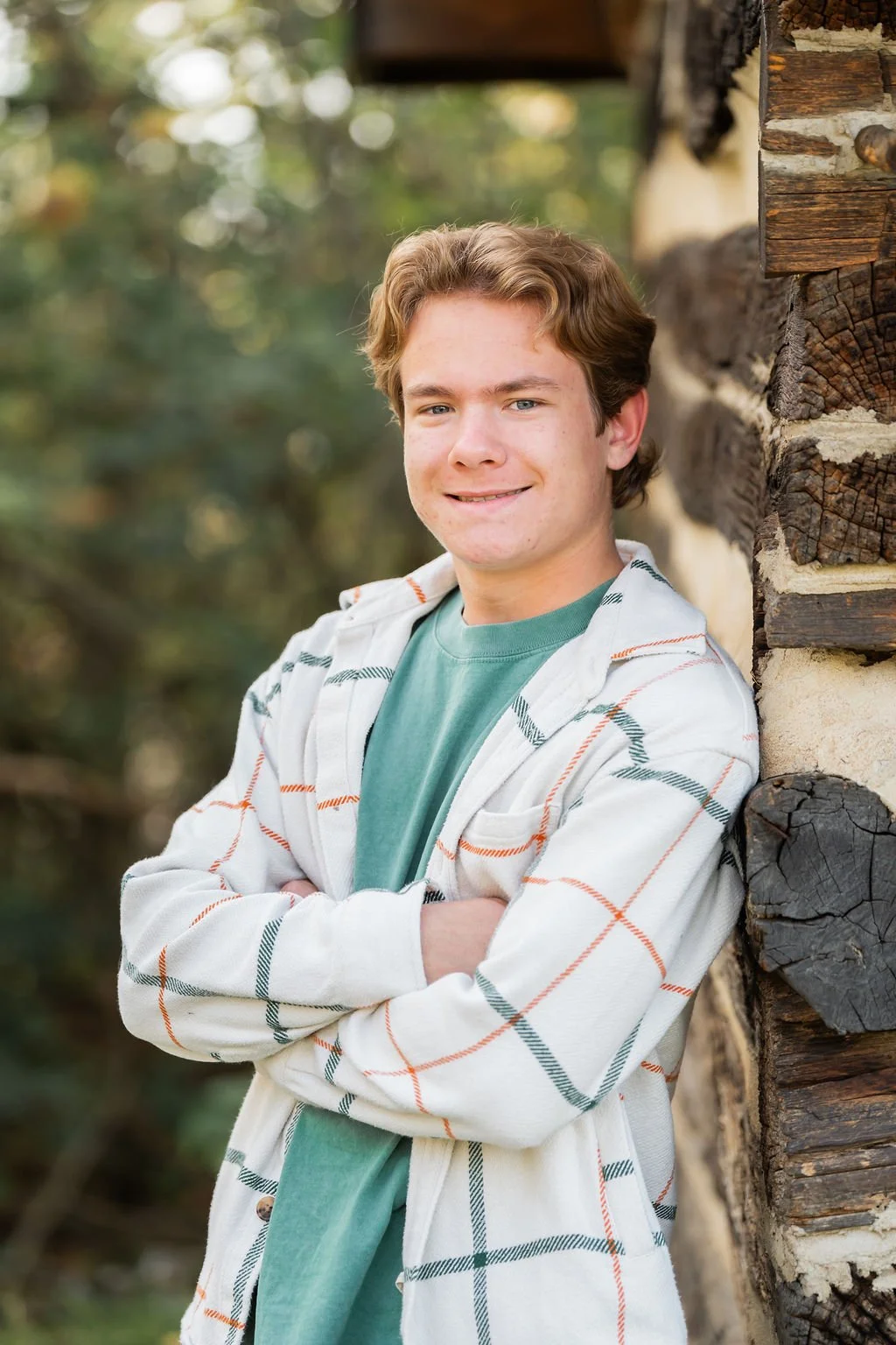 Young man with wavy brown hair smiling, standing outdoors next to a log cabin wall with arms crossed, wearing a light-colored plaid jacket over a green shirt.
