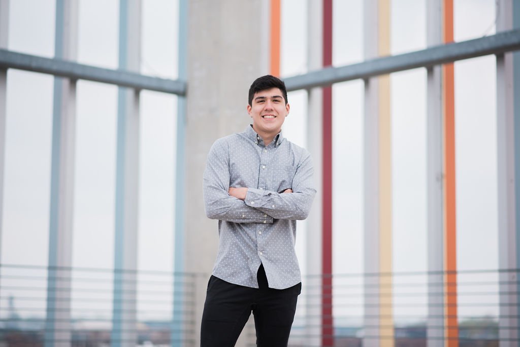 A young man smiling with arms crossed standing in front of a modern architectural background with colorful vertical beams.