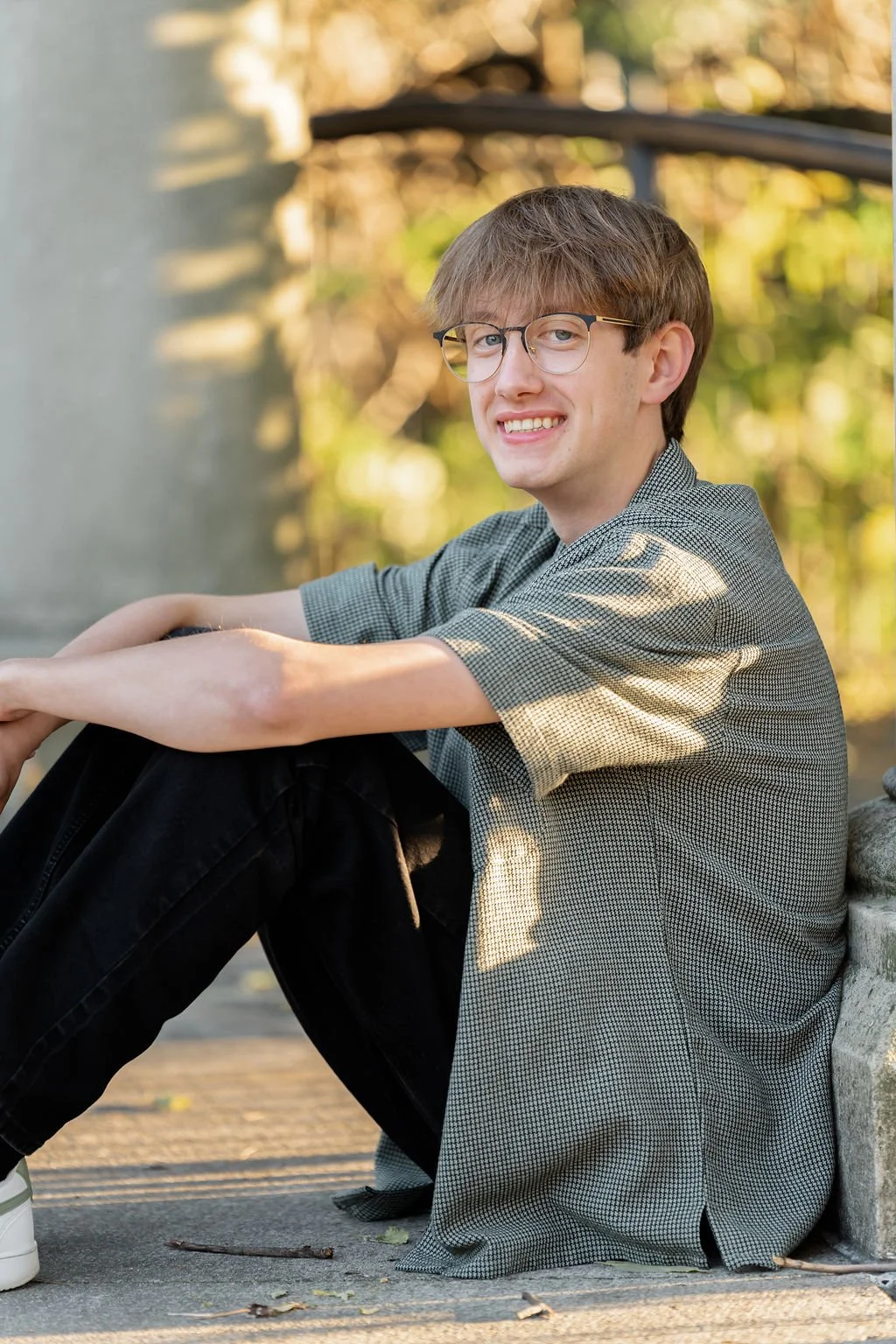 A young man with glasses and brown hair sitting outside on a concrete surface, smiling at the camera, with trees and sunlight in the background.