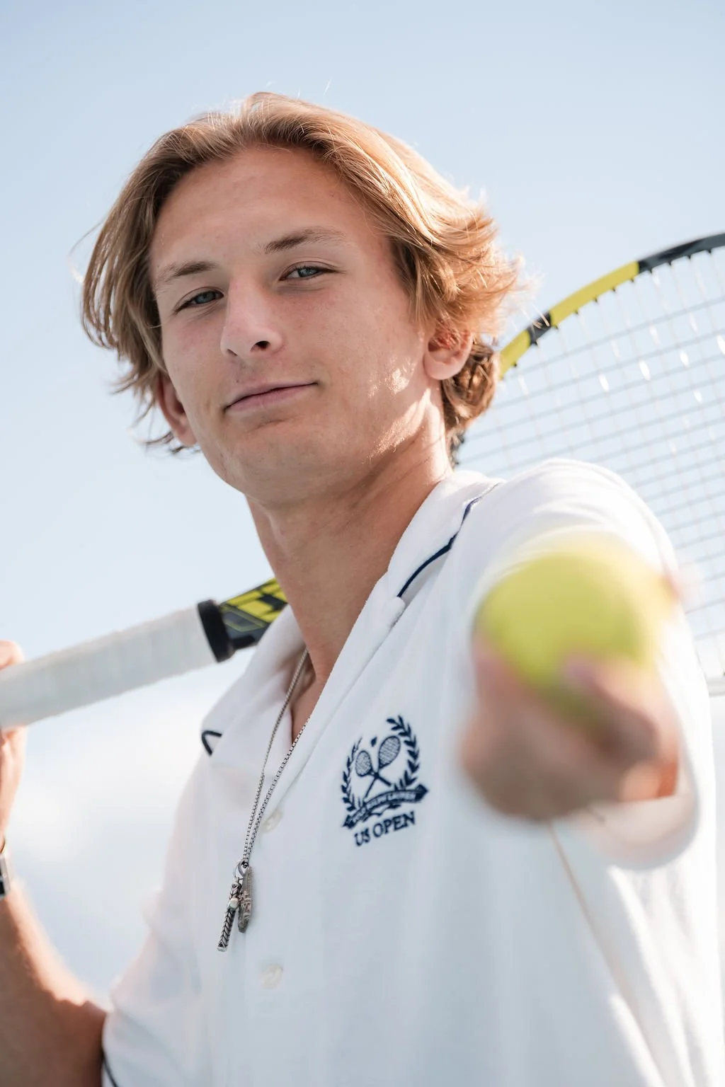 A young man with light brown hair holding a tennis racket over his shoulder, wearing a white U.S. Open polo shirt with a tennis emblem, standing outdoors under a clear sky.