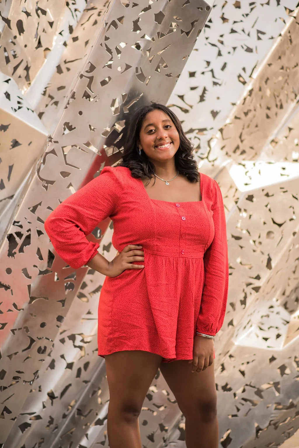 A woman with medium skin tone, black hair, and wearing a red dress with long sleeves, stands in front of a metallic patterned wall, smiling at the camera.
