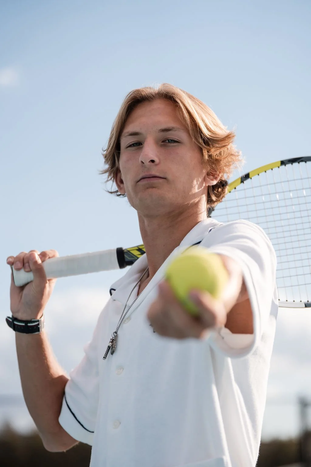 Young man with wavy light brown hair holding tennis racket over his right shoulder and a tennis ball extended towards the camera, wearing a white polo shirt against a clear blue sky.