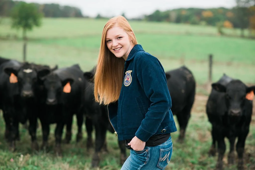A young woman with long red hair smiling in front of a group of black cows on a green farm with trees in the background.