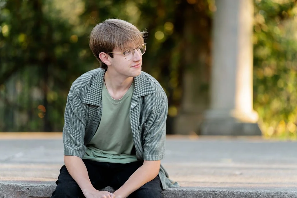 A young man with glasses and light brown hair sitting outdoors on a stone step, with a blurred background of trees and a column.