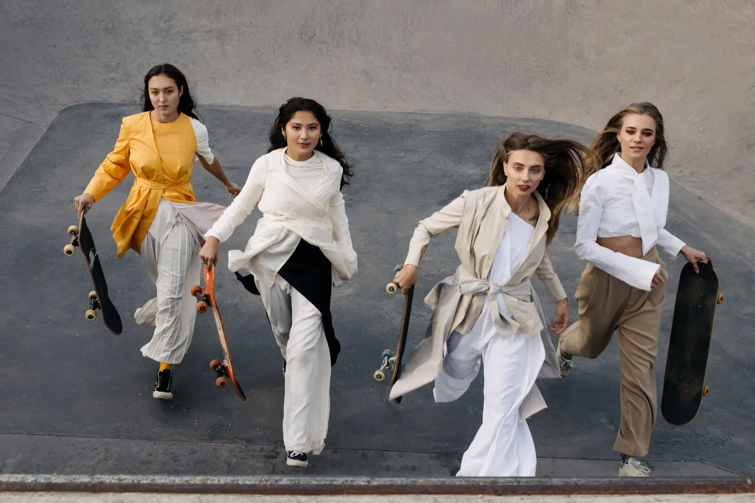 Five women walking in a skate park holding skateboards, dressed in fashion-forward casual outfits.