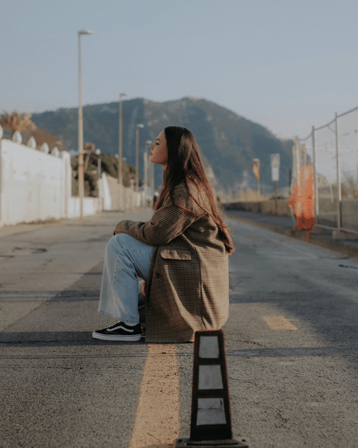 Young woman sitting on the road during daytime, wearing a long brown coat, light blue jeans, and black sneakers, with a mountain in the background and a traffic cone in the foreground.