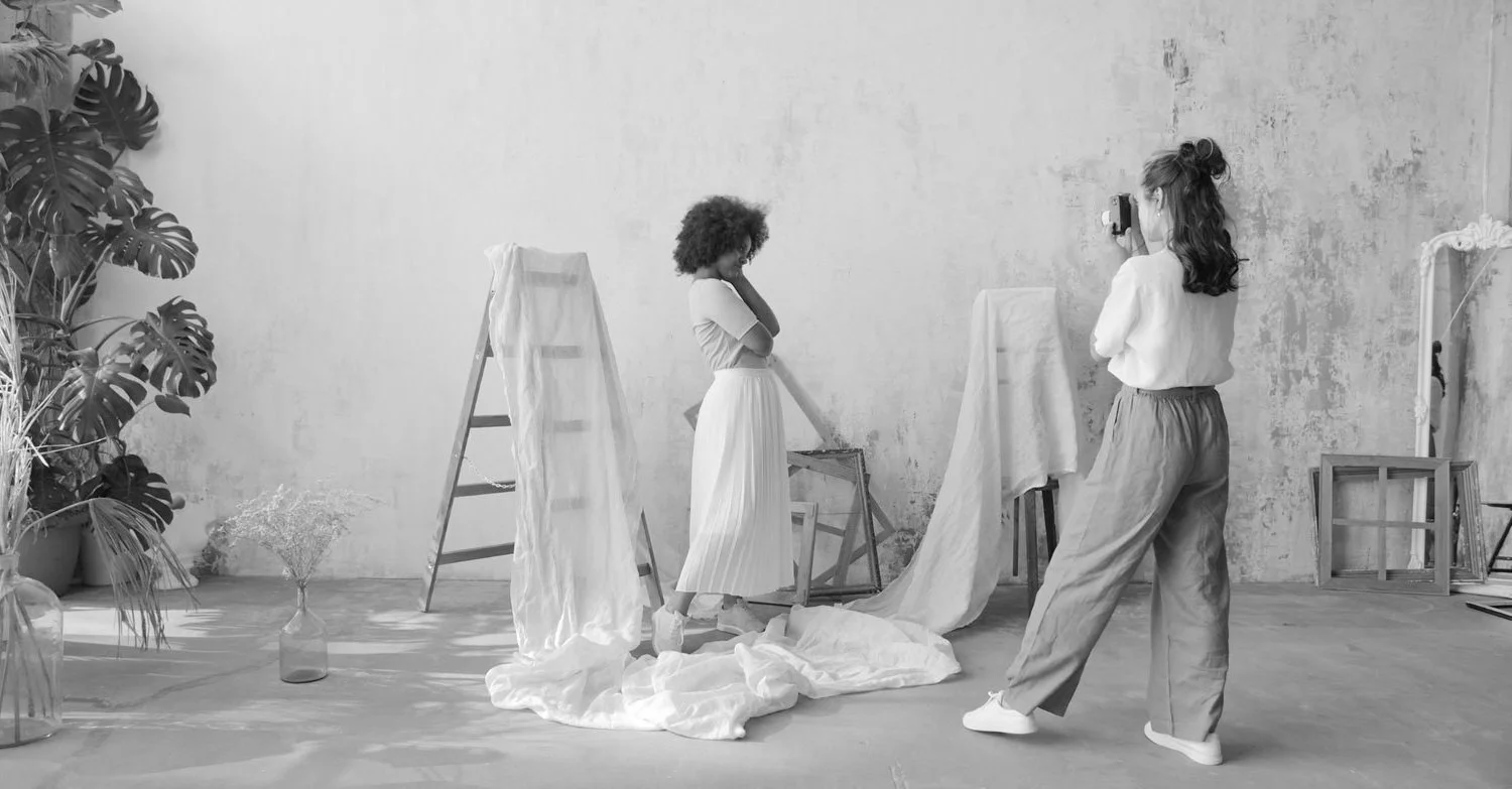 Two women in a photography studio, one posing and the other taking a picture, with ladders, fabric draped over furniture, and plants in the background, all in black and white.
