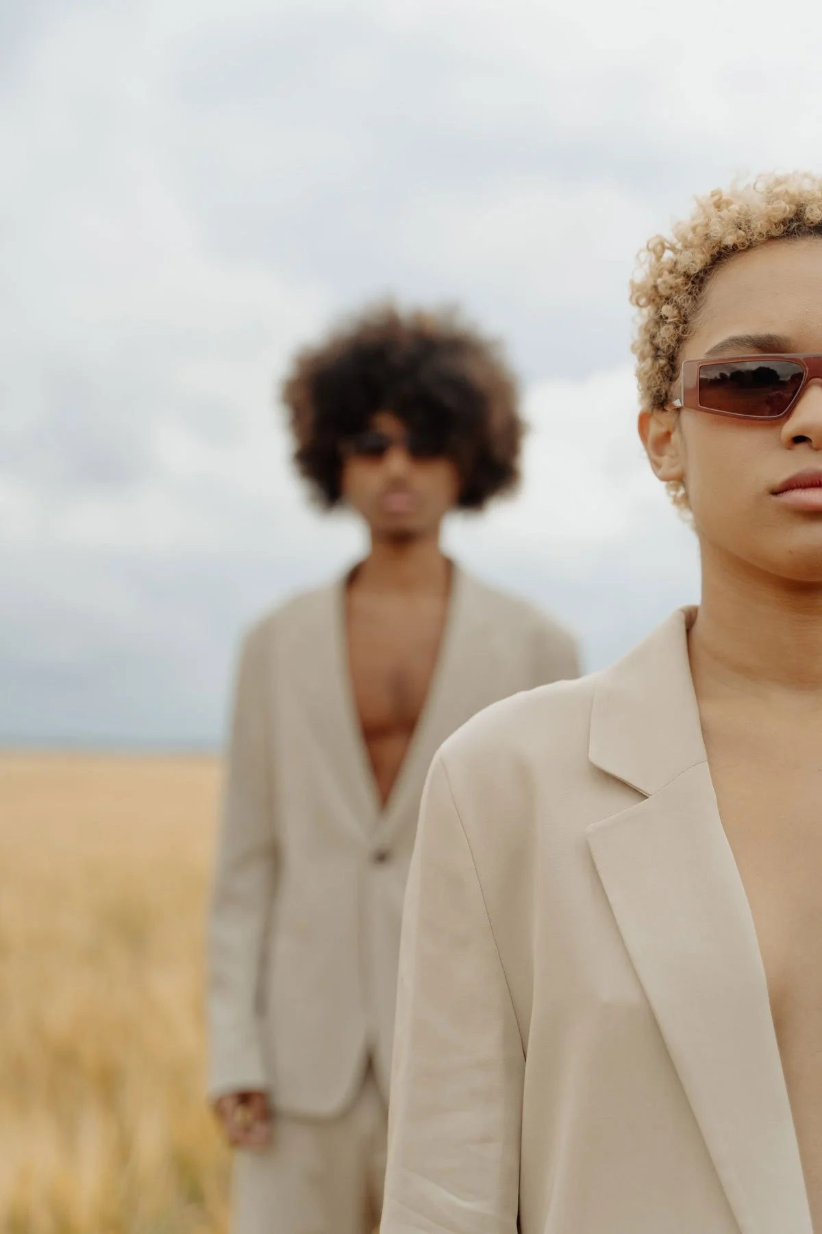 Two women in beige suits standing outdoors in a field with cloudy sky in the background. One woman is in the foreground wearing sunglasses, while the other is in the background, slightly out of focus.