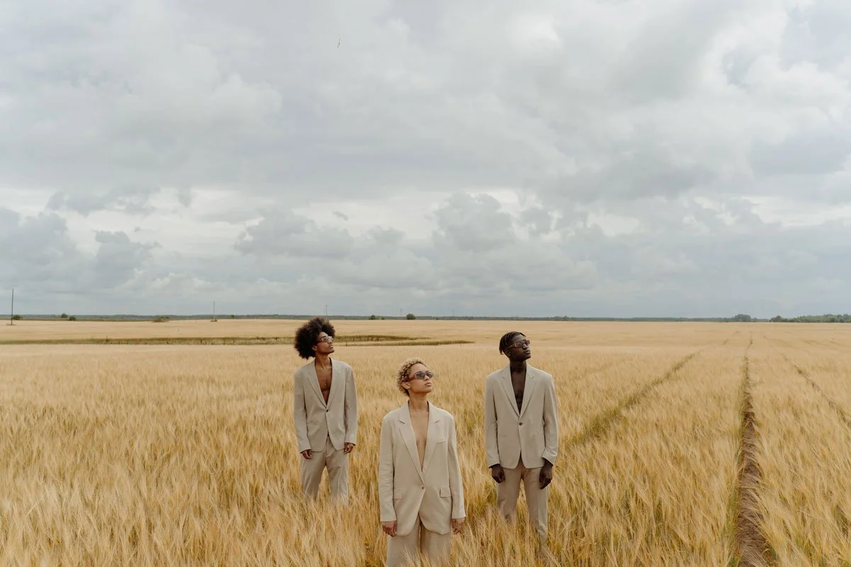 Three women in beige suits stand in a wheat field under a cloudy sky, gazing upwards.