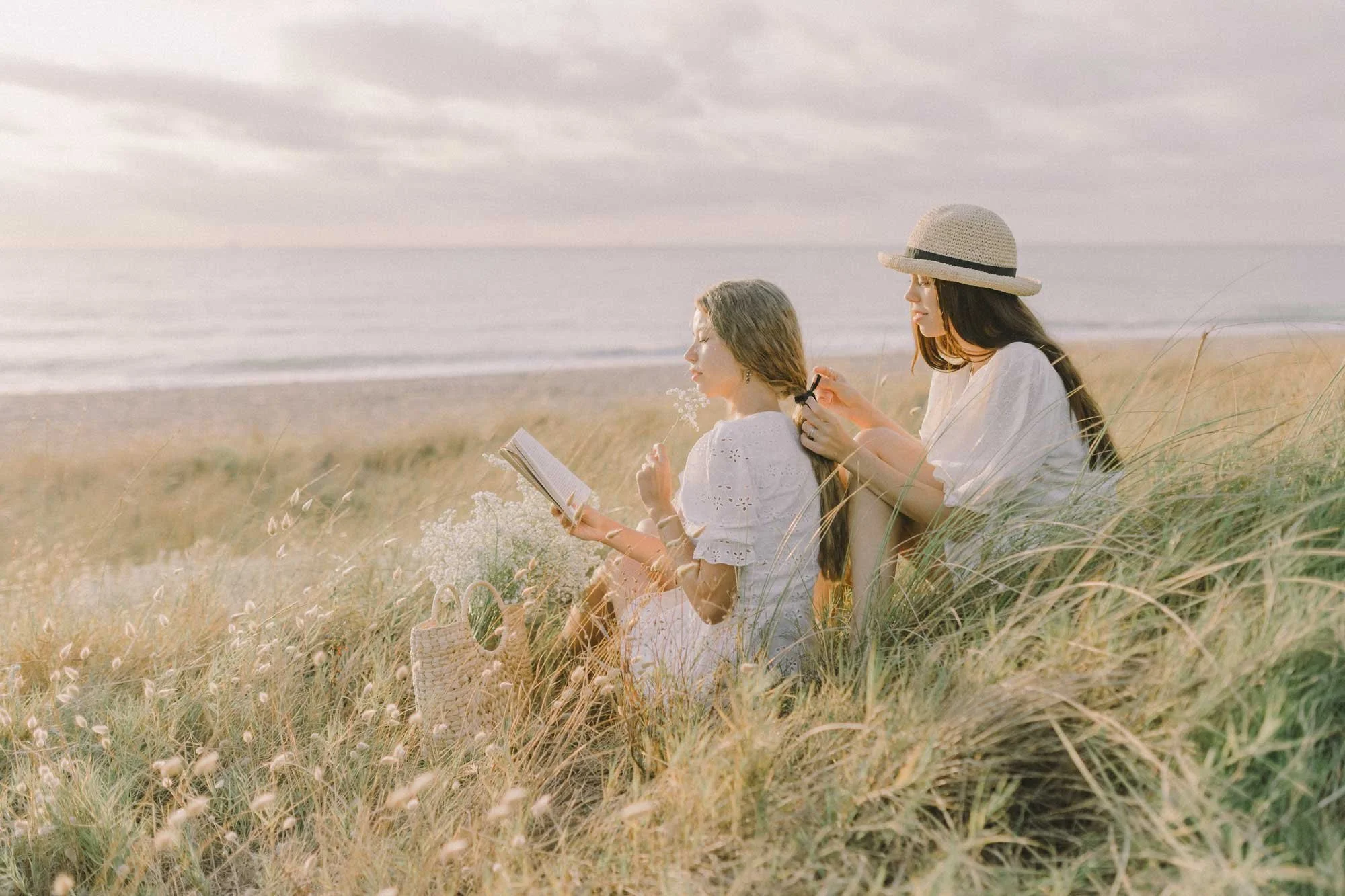 Two women sitting in tall grass on a beach, one reading a book and the other sitting behind applying makeup, with a beach and ocean in the background.