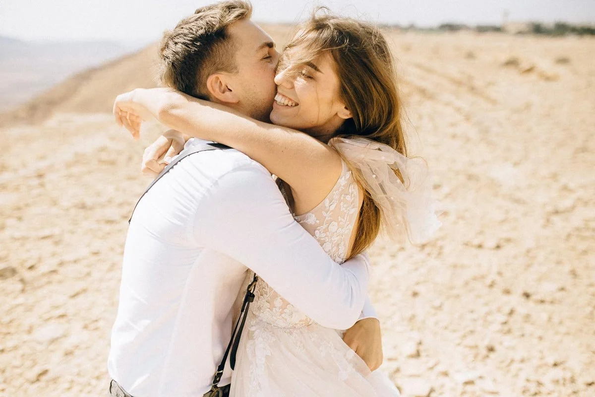 A young man and woman embrace and smile on a sandy beach, with the man kissing the woman's cheek.