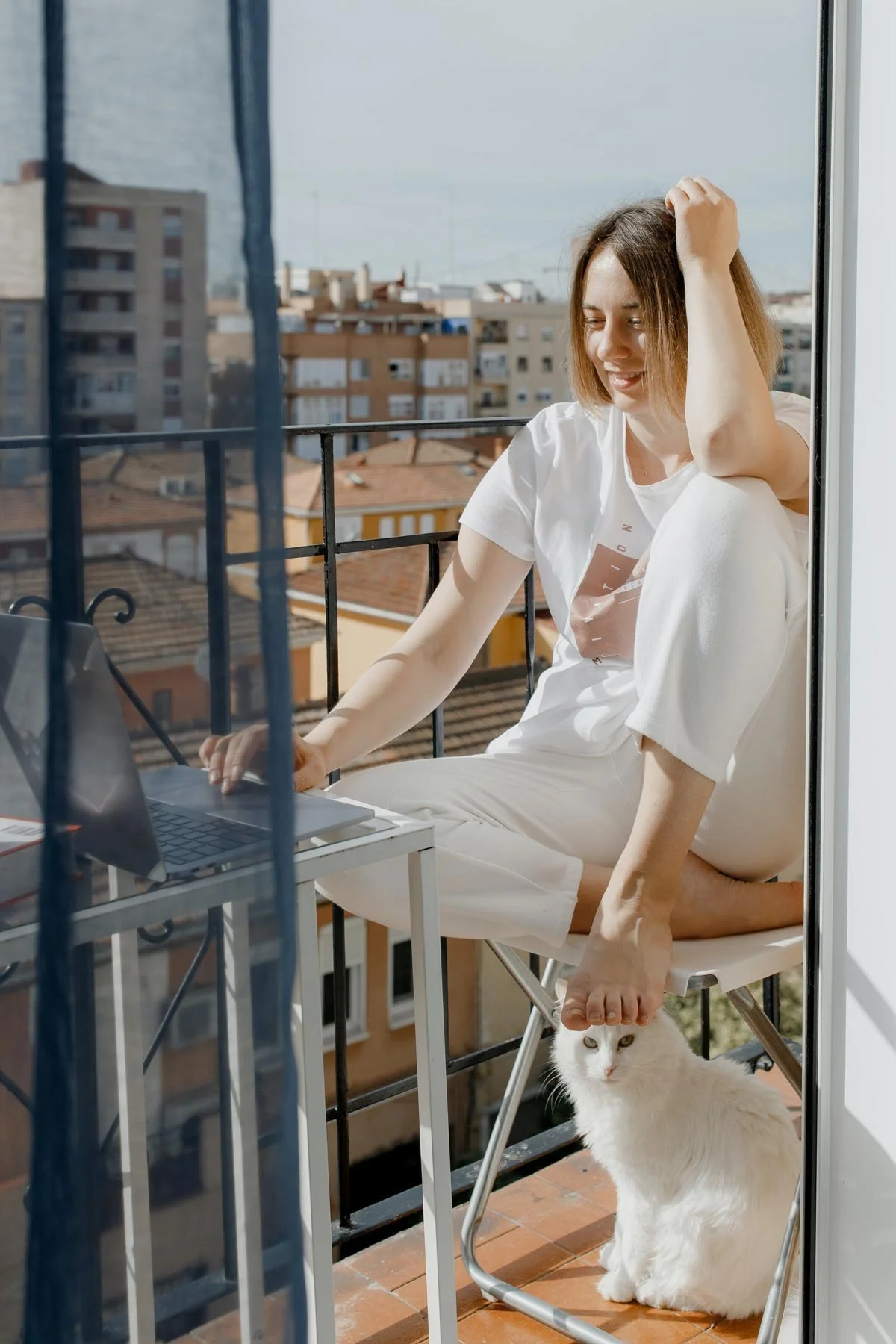 A woman sitting on a balcony with a laptop, petting a white cat under her foot, with buildings and a cloudy sky in the background.