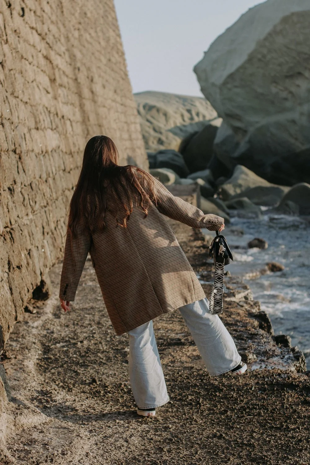 A woman with long brown hair wearing a checkered coat and white pants walking along a rocky beach near a stone wall with large rocks and the ocean in the background.
