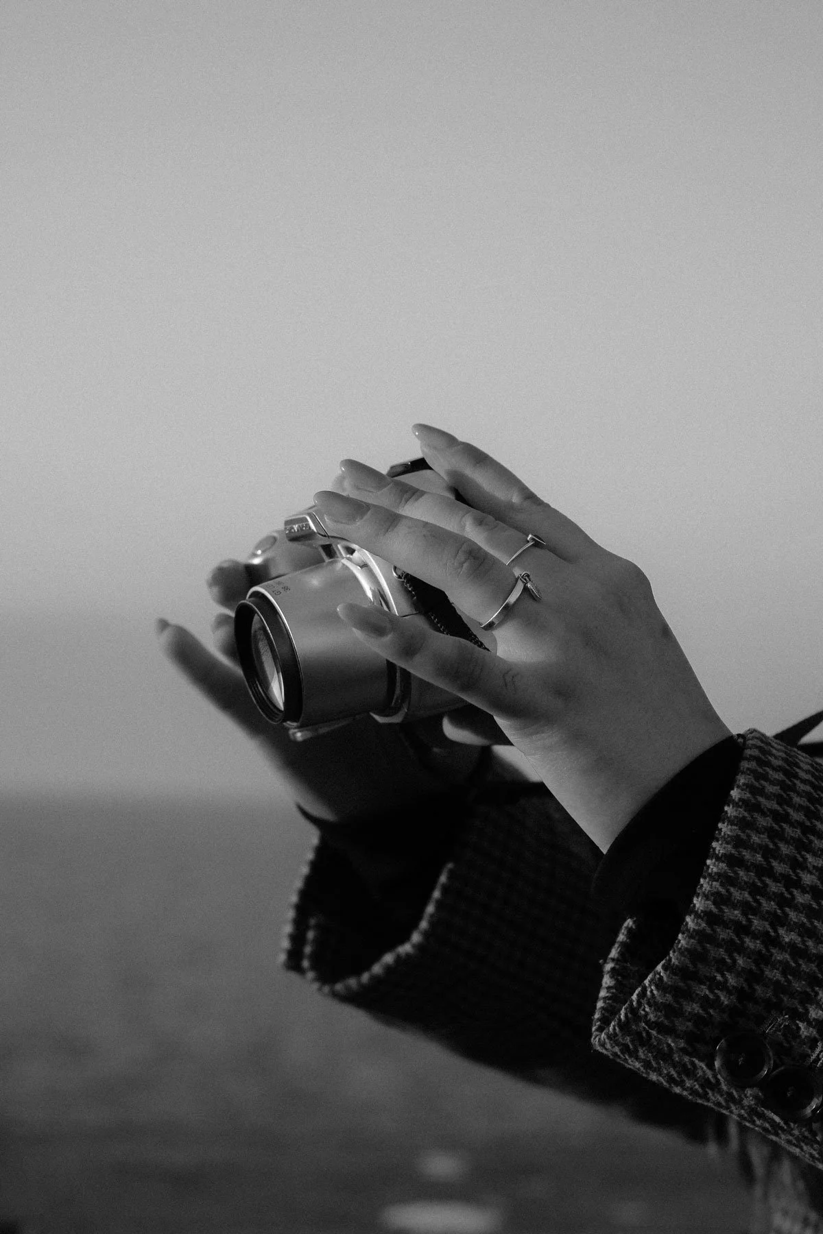 Close-up of a person’s hand holding a vintage camera, wearing rings and a patterned coat, in black and white.
