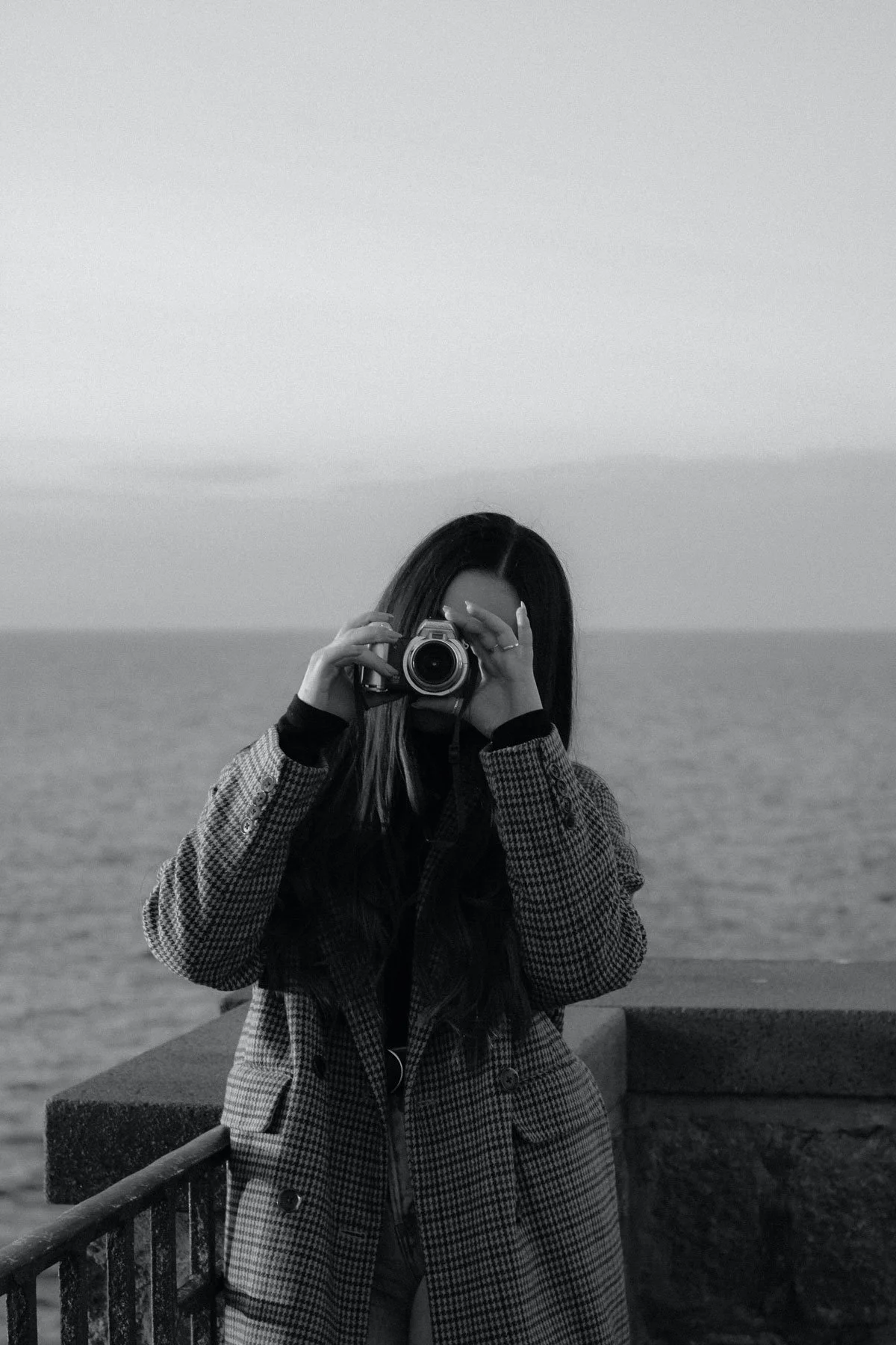 A woman with long dark hair wearing a houndstooth coat, taking a photo with a camera near a body of water, in black and white.