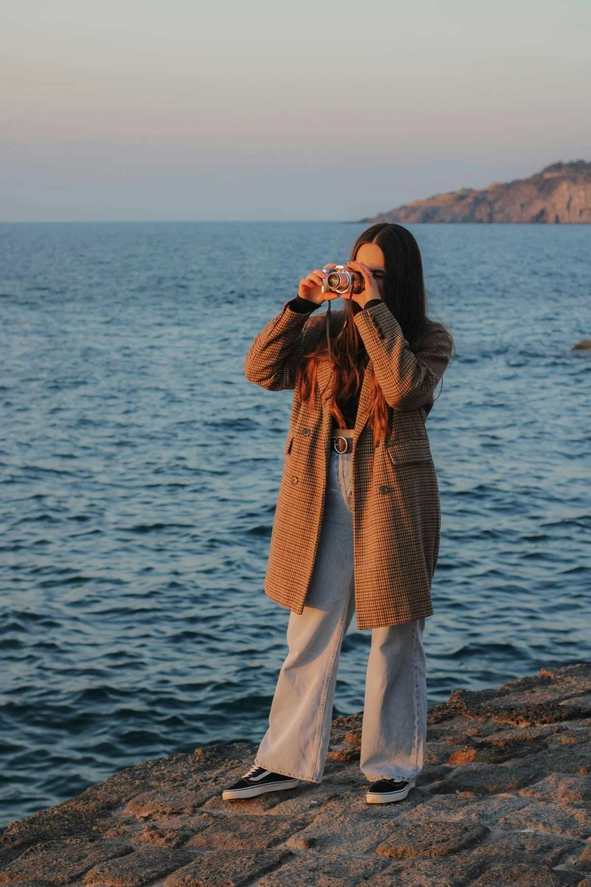 A woman standing on a rocky shoreline taking a photo with a camera, overlooking the ocean at sunset.
