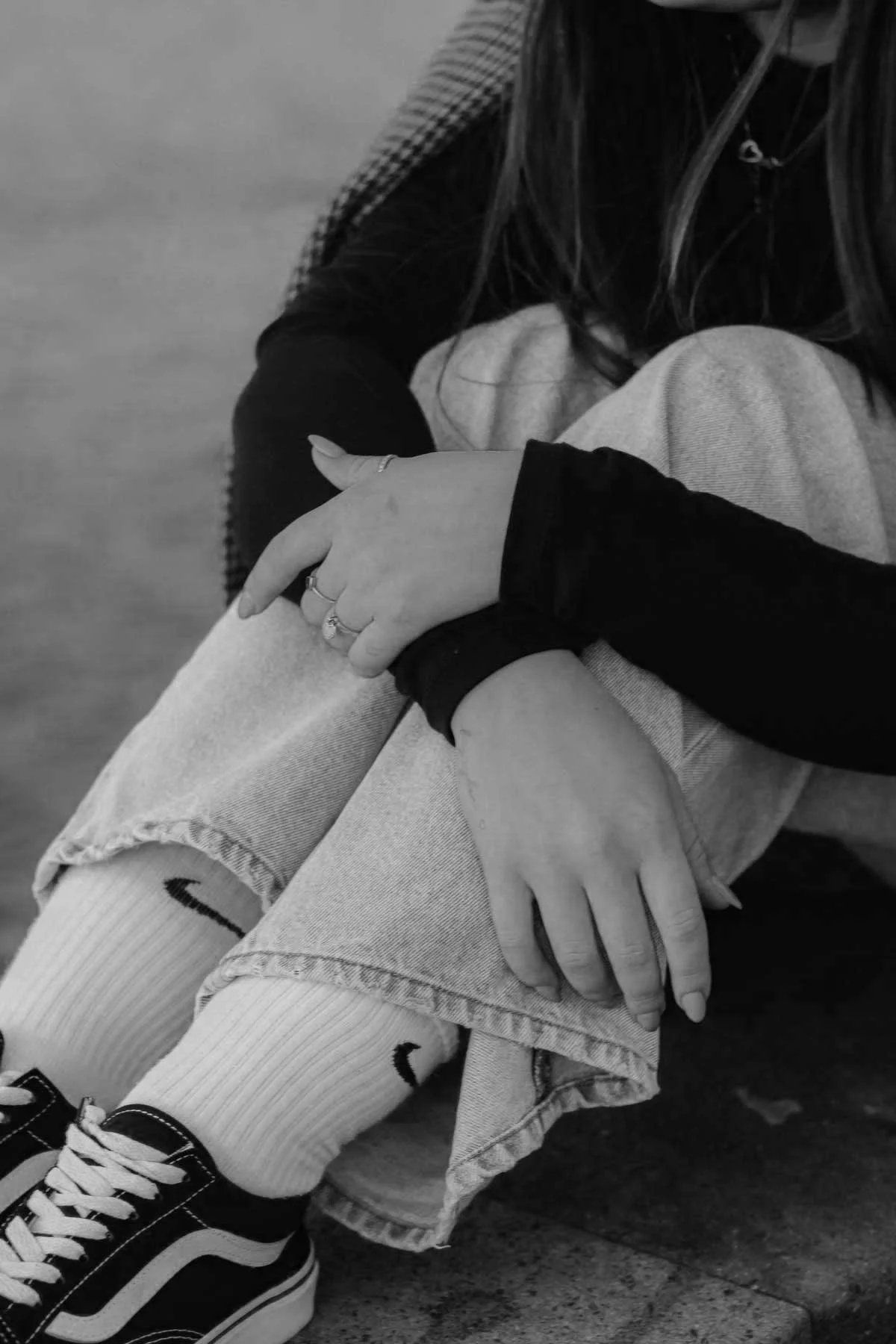 A black and white photo of a person sitting on the ground with their arms around their knees. They are wearing sneakers, knee-high socks, ripped jeans, and a long-sleeve shirt.