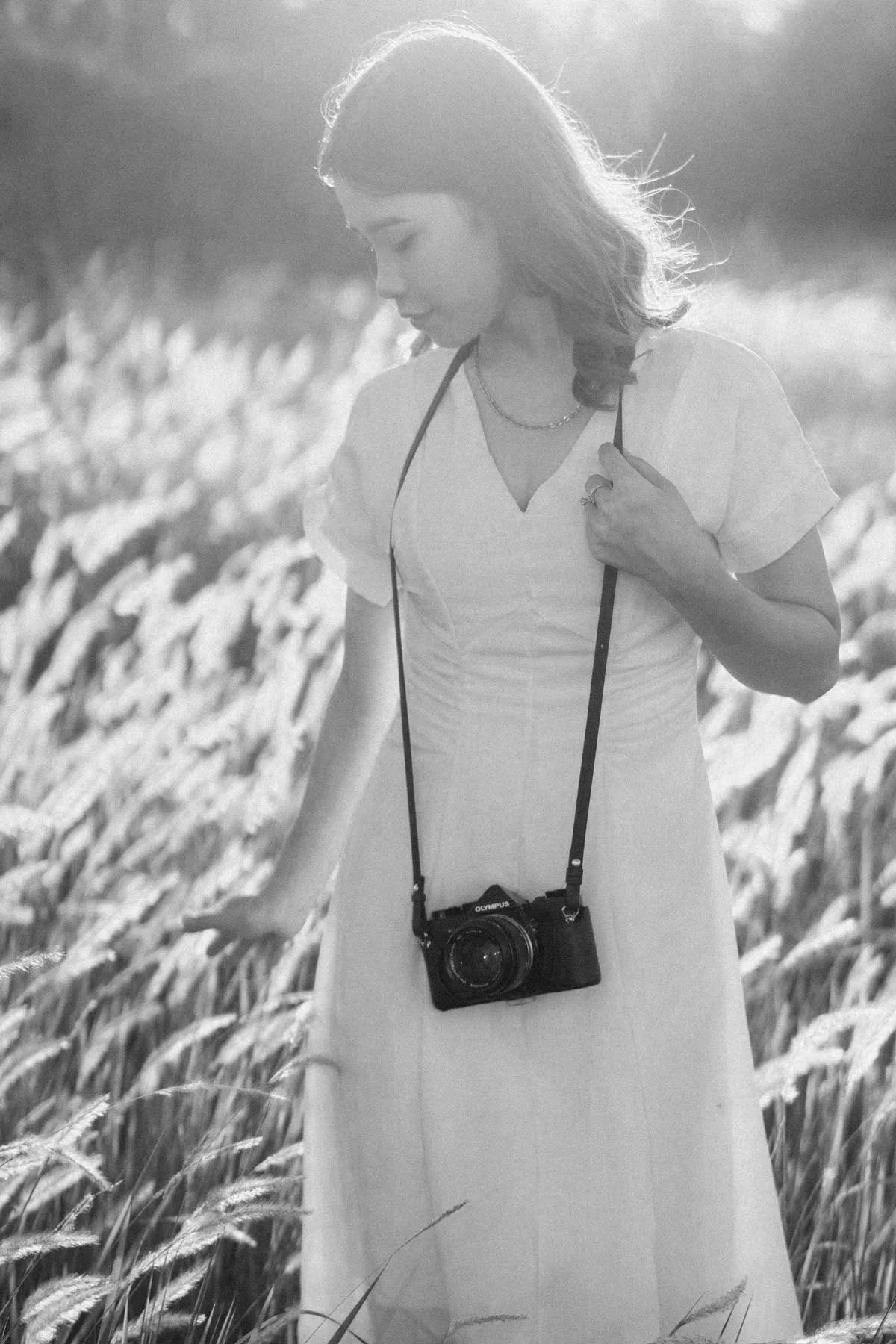 A woman in a dress standing in a field of tall grass, holding a camera hanging from her neck, with sunlight illuminating her hair.