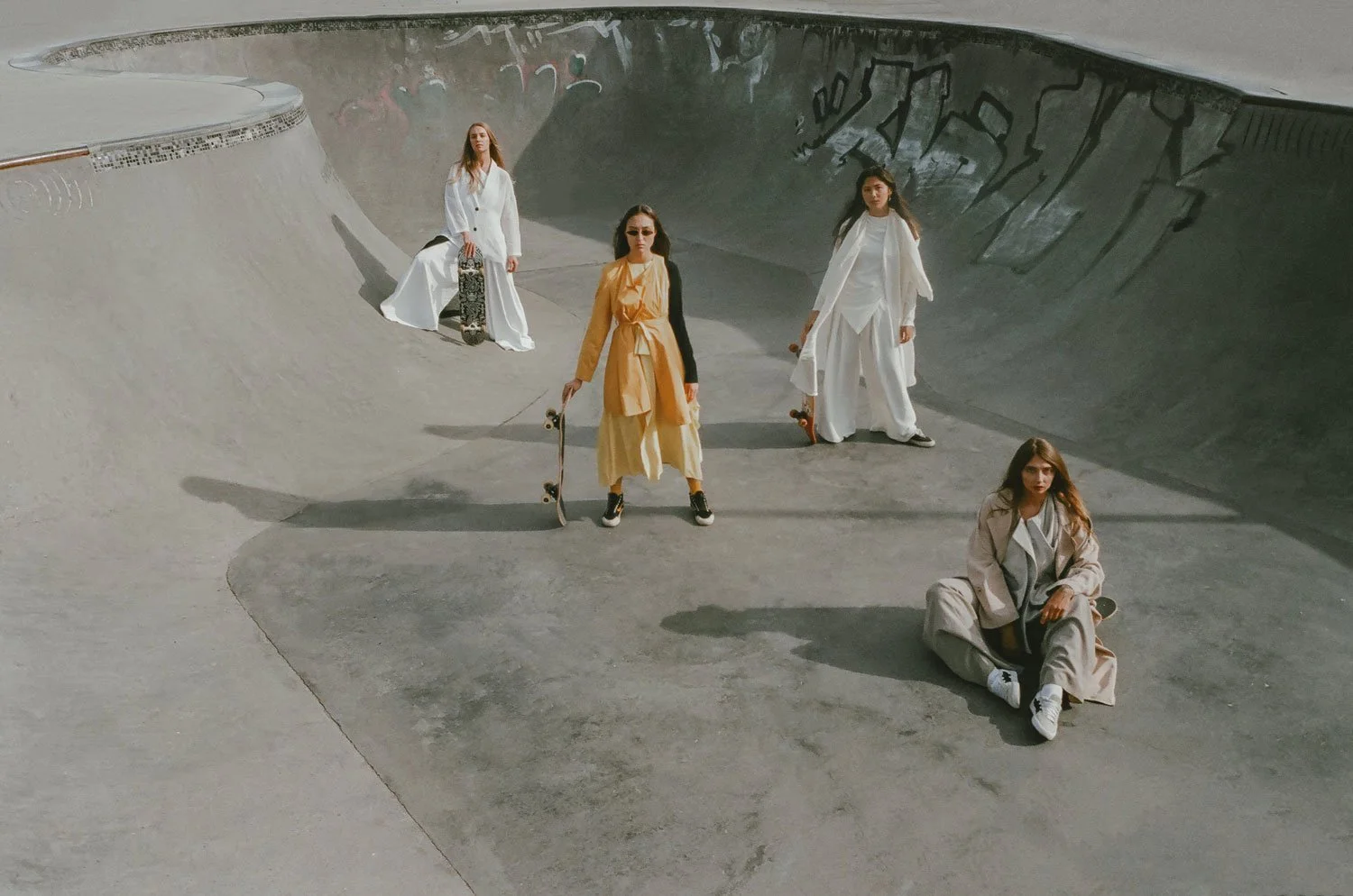Five women in fashionable clothing at a skatepark with concrete ramps and graffiti, some holding skateboards, posing for a photo.