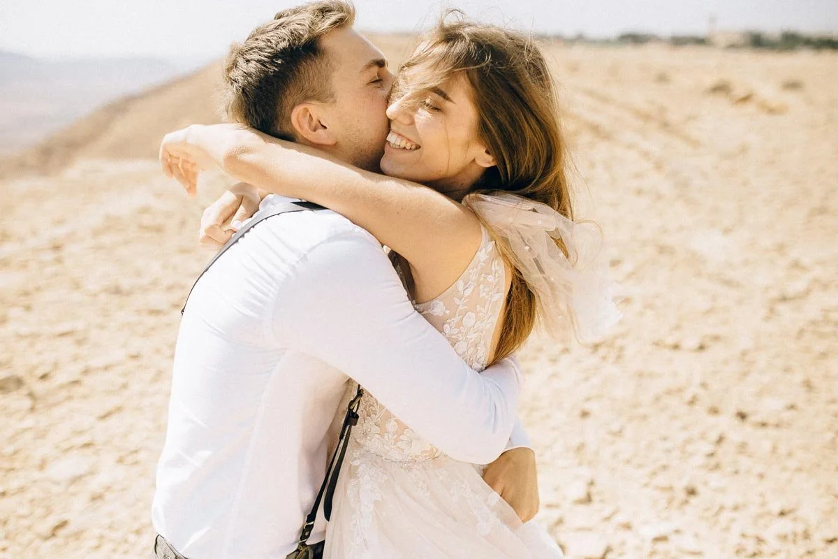 A happy couple embracing and smiling on a sandy beach, with the woman leaning into the man's shoulder as they share an affectionate hug.