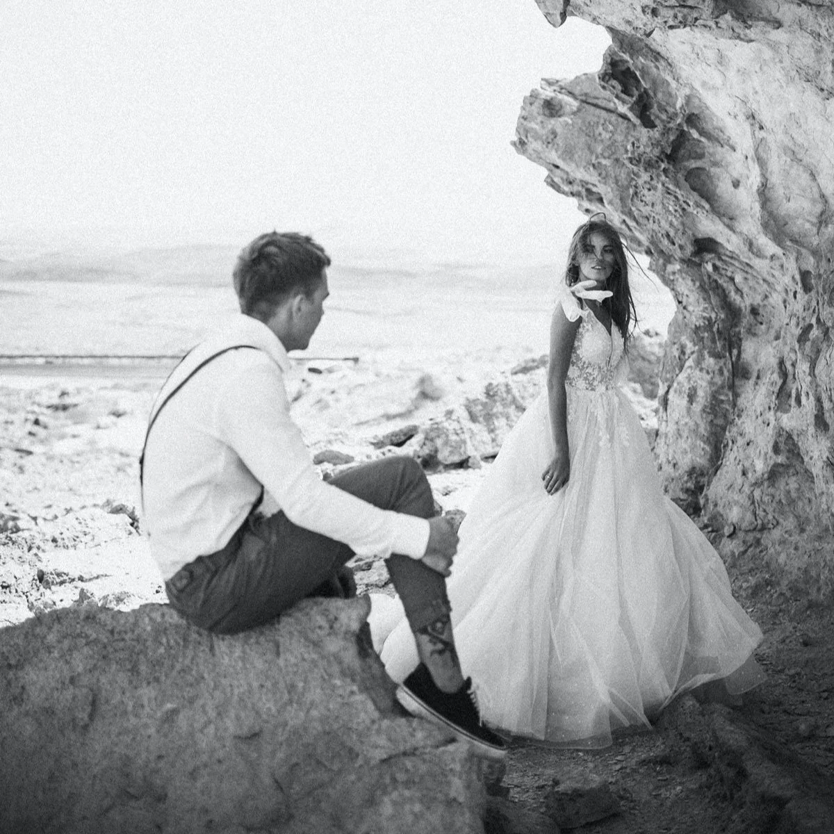 A black and white photo of a man and a woman on a rocky beach. The man is sitting on a rock, looking at the woman, who is standing next to a large rock formation. The woman is wearing a flowing wedding dress and smiling at the camera.