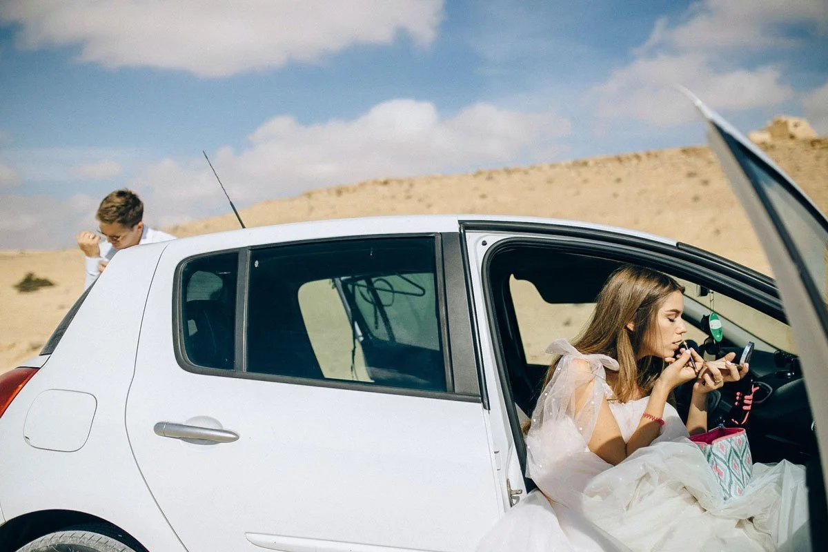 A woman in a white dress sitting inside a white car, applying makeup or makeup remover while looking at her phone, in a desert landscape with blue sky and clouds.