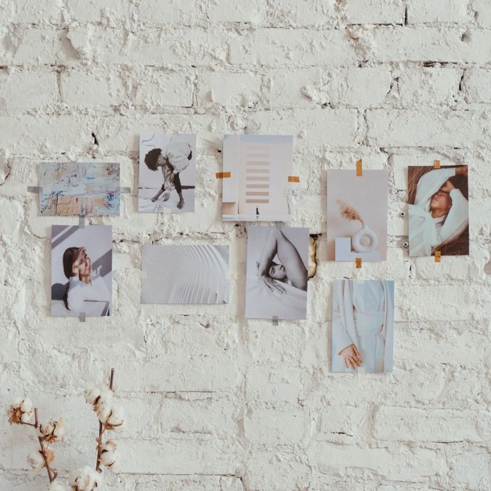 A white brick wall with nine photographs and artwork taped to it, including images of people, abstract art, and architectural visuals. There is also a cotton branch in the lower left corner.