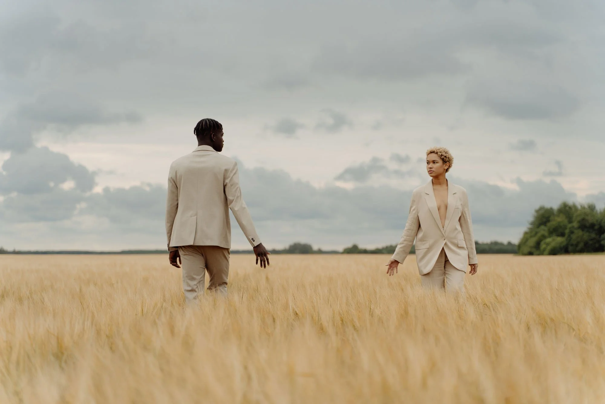 Two people in beige suits walking through a golden wheat field under a cloudy sky.