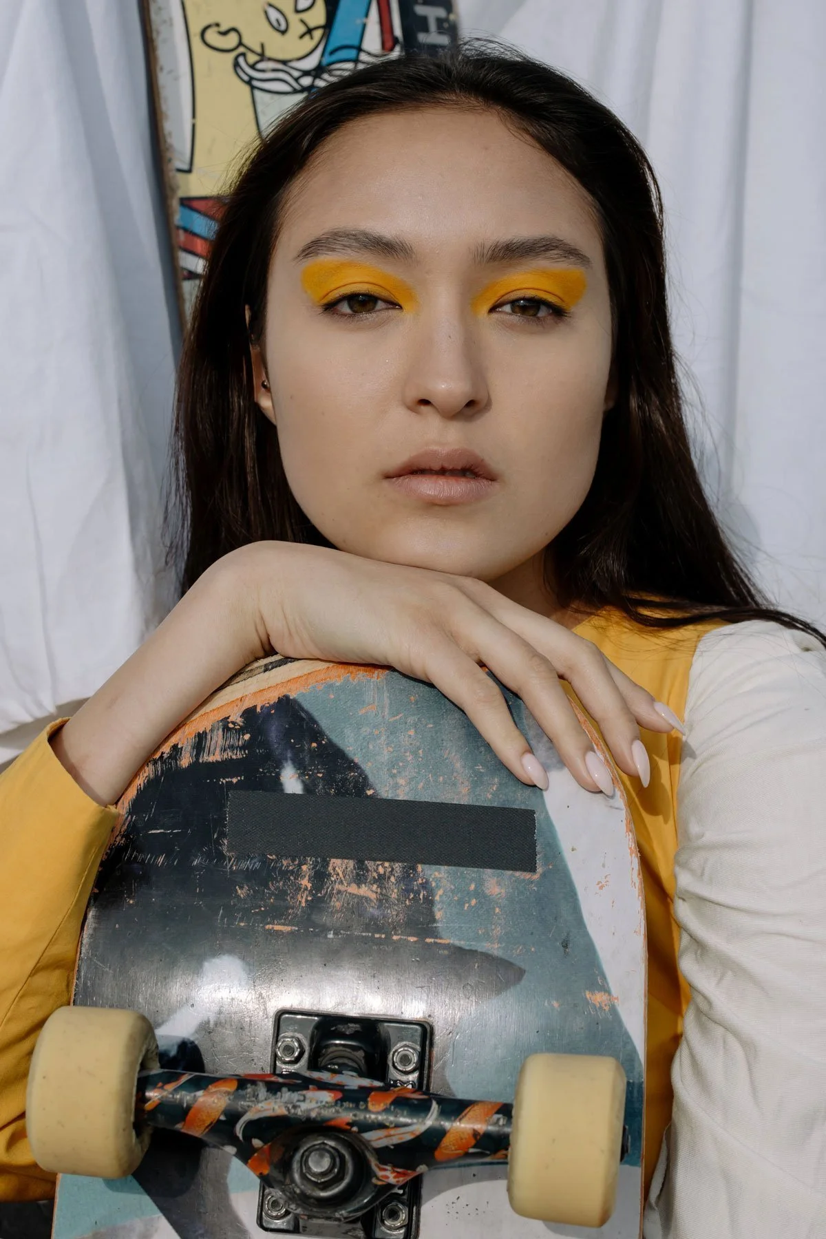 A woman with yellow eyeshadow leaning on a skateboard that has worn-out paint and yellow wheels, with a white curtain and a cartoon art background behind her.