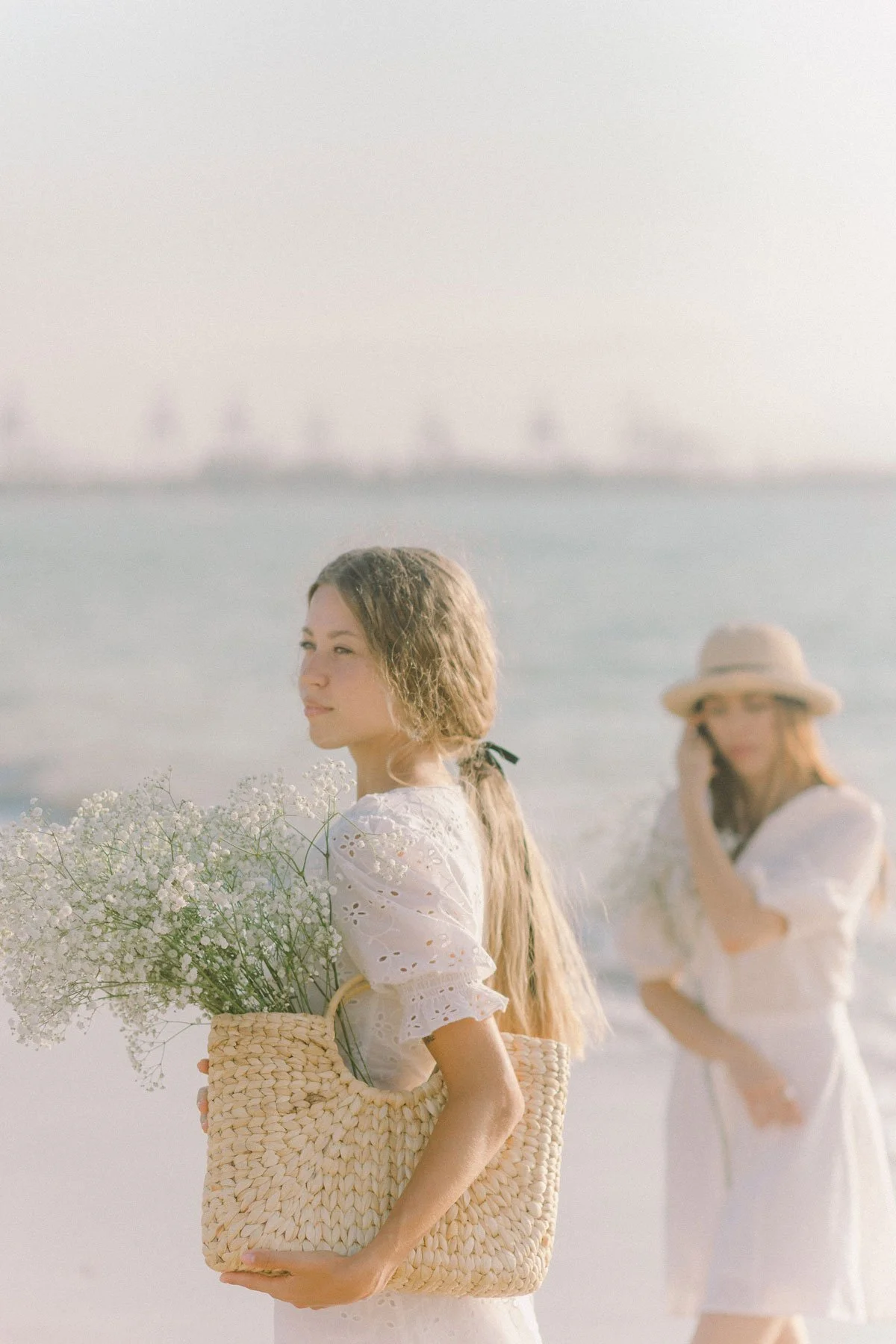 Two women standing on the beach, wearing white dresses, one holding a basket with white flowers, and the other talking on the phone, with a blurred background of water and sailboats.