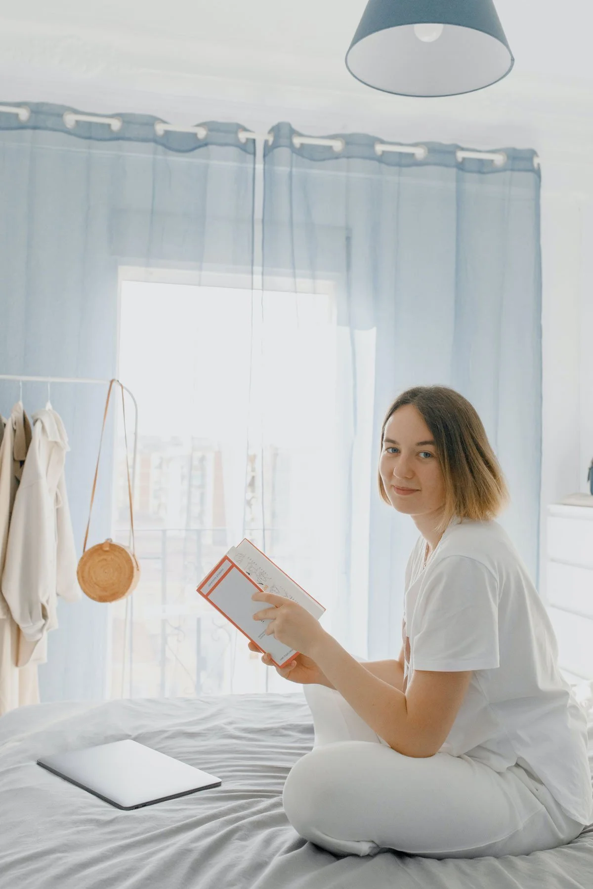 A young woman with short brown hair sitting on a bed in a bright bedroom, holding a book and looking at the camera, with a laptop on the bed, soft sunlight coming through the window, and light blue curtains.