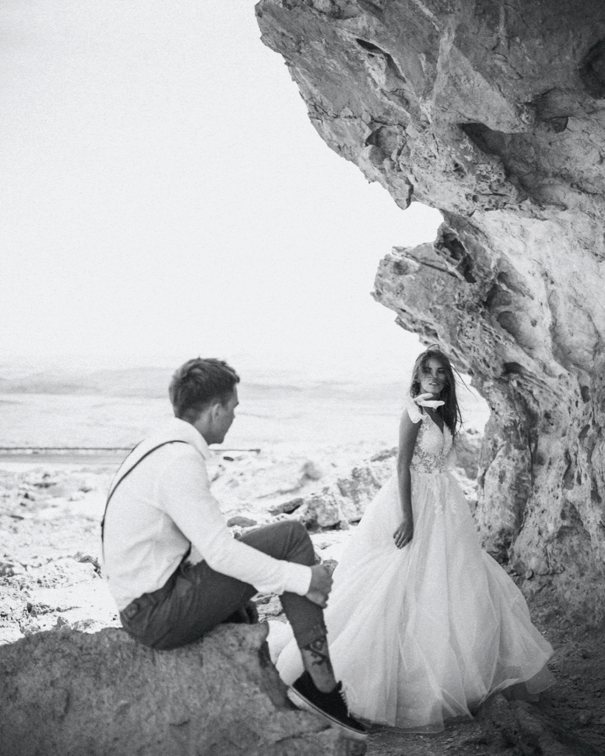 A black and white photograph of a woman in a wedding dress standing near a rocky formation on a beach, with a man sitting on a rock nearby.