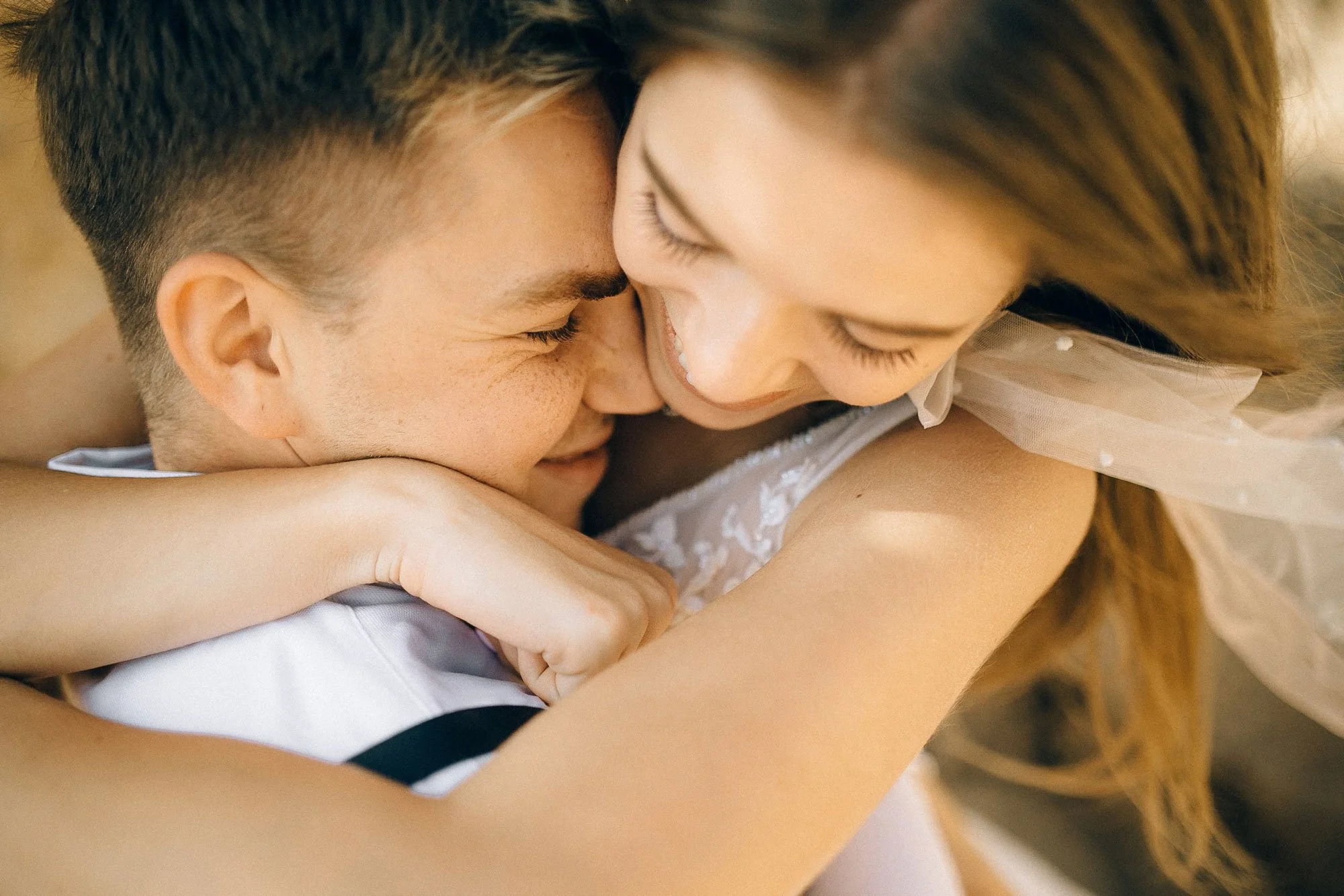 A close-up of a couple hugging, smiling, with their faces close together and eyes closed, showing affection and intimacy.