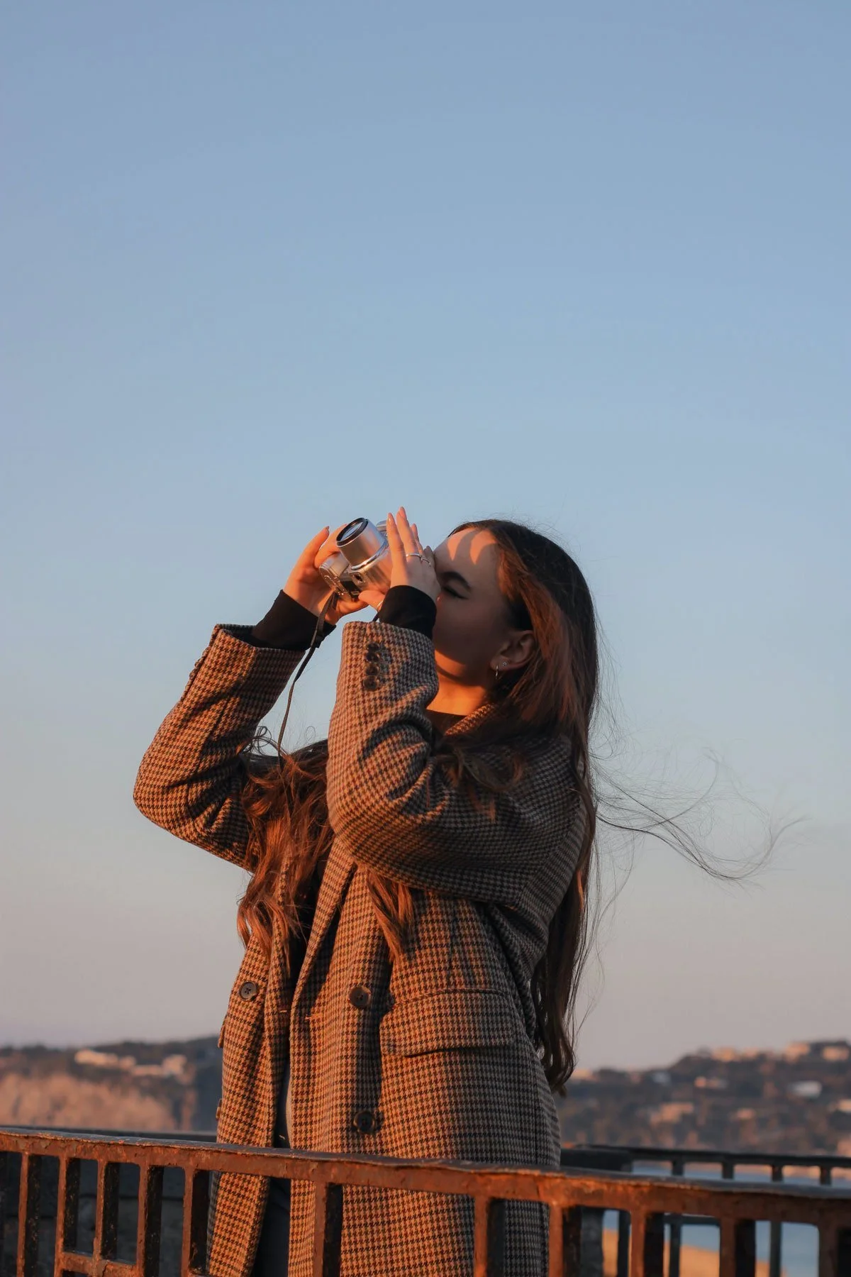 A woman with long hair wearing a houndstooth coat taking a photo with a camera outdoors during sunset.
