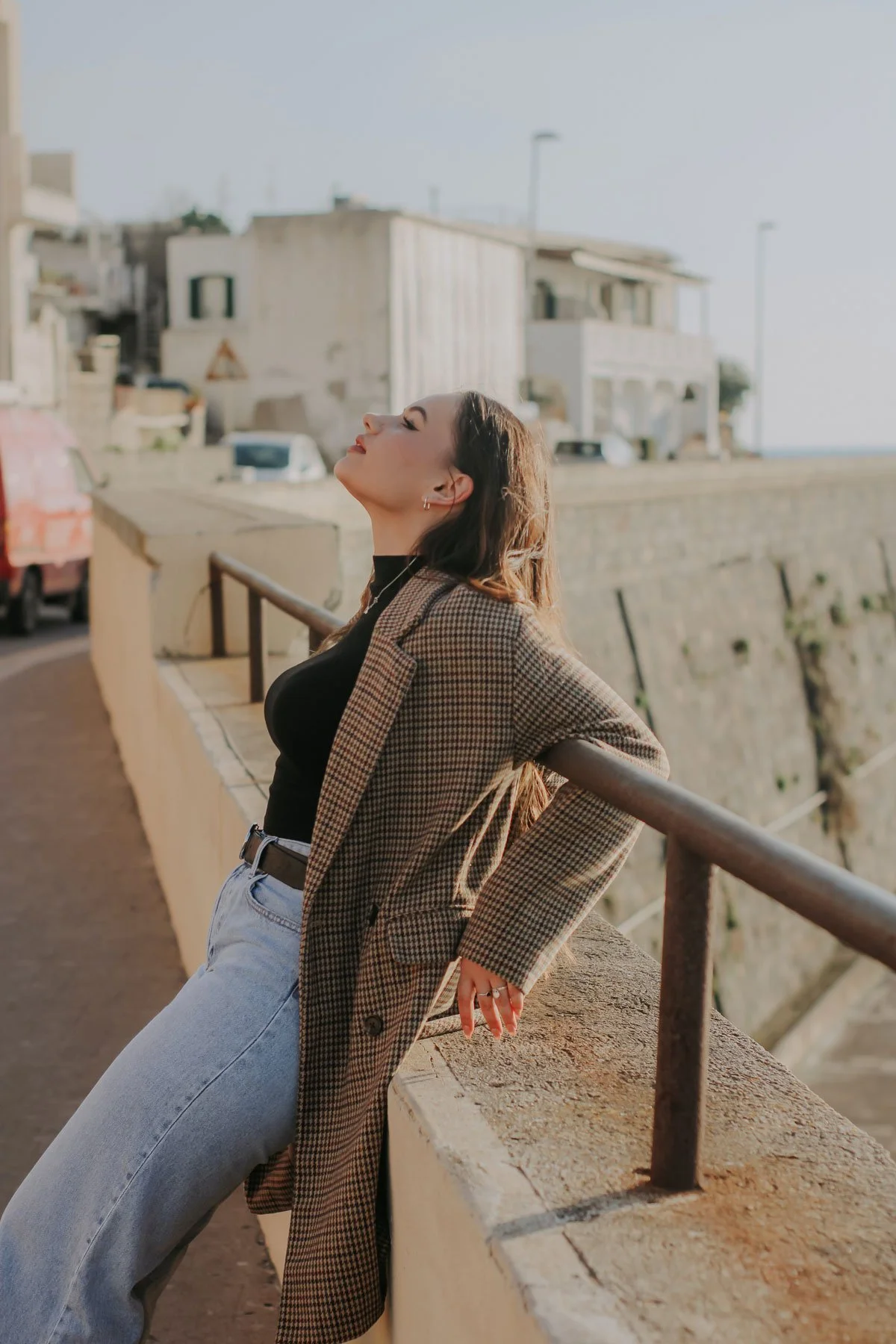 Woman with long hair leaning against a railing with her eyes closed, wearing a checkered blazer and black top, outdoors in an urban setting during daylight.