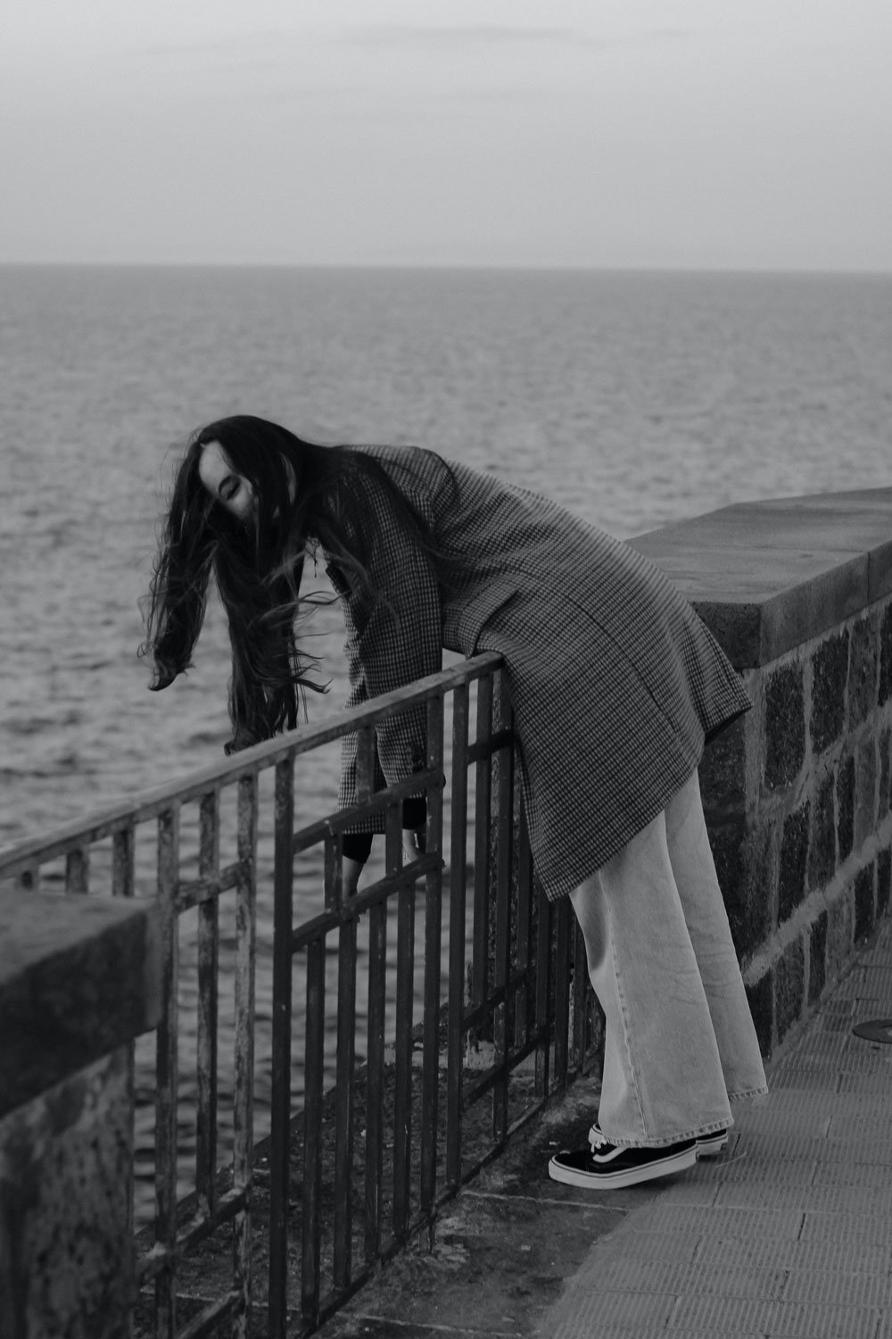 A woman leaning over a metal railing on a seaside promenade, looking into the water, with long hair flowing, dressed in a checkered coat, light trousers, and sneakers.