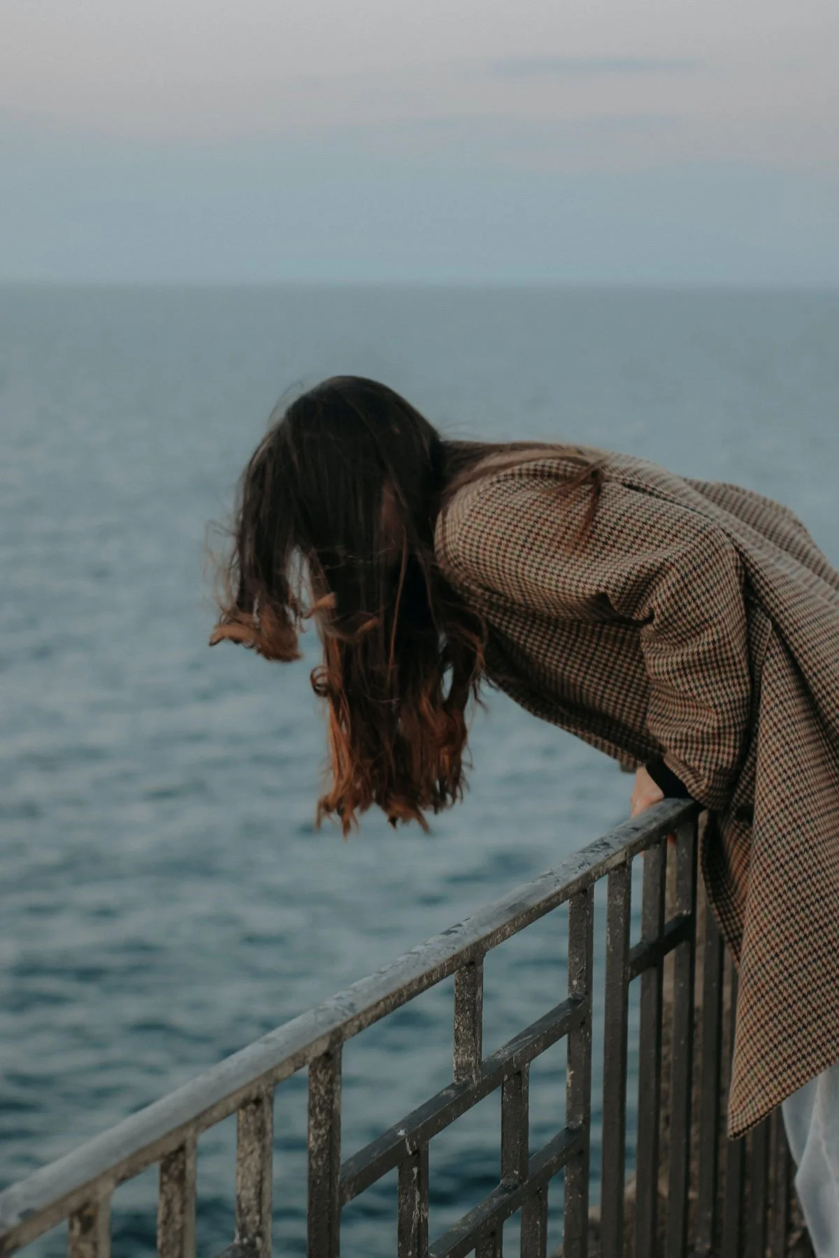A woman with long brown hair leaning over a metal railing, looking down at the water in an ocean or large body of water, with an overcast sky in the background.