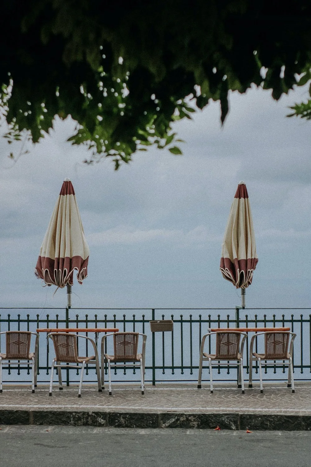Empty outdoor seating area with five chairs, two closed umbrellas, overlooking the sea under cloudy sky, partially covered by tree branches.