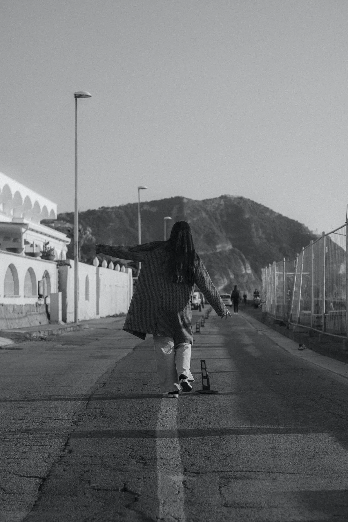 A person with long hair walking down a street with arms outstretched, walking away from the camera. The street is lined with buildings on the left and a fenced area on the right, with mountains in the background. The image is in black and white.