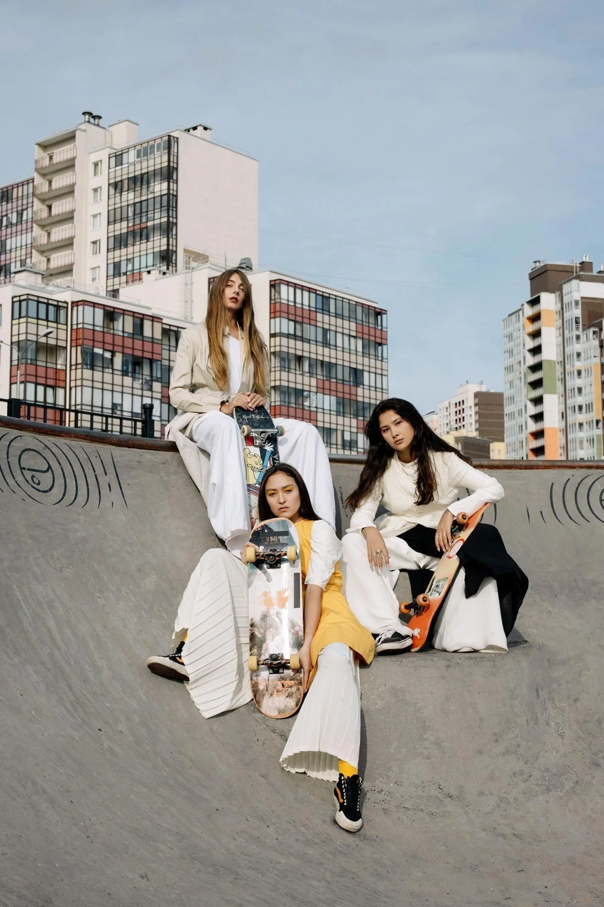 Four young women at a skate park with skating boards, surrounded by modern multi-story apartment buildings, under a partly cloudy sky.