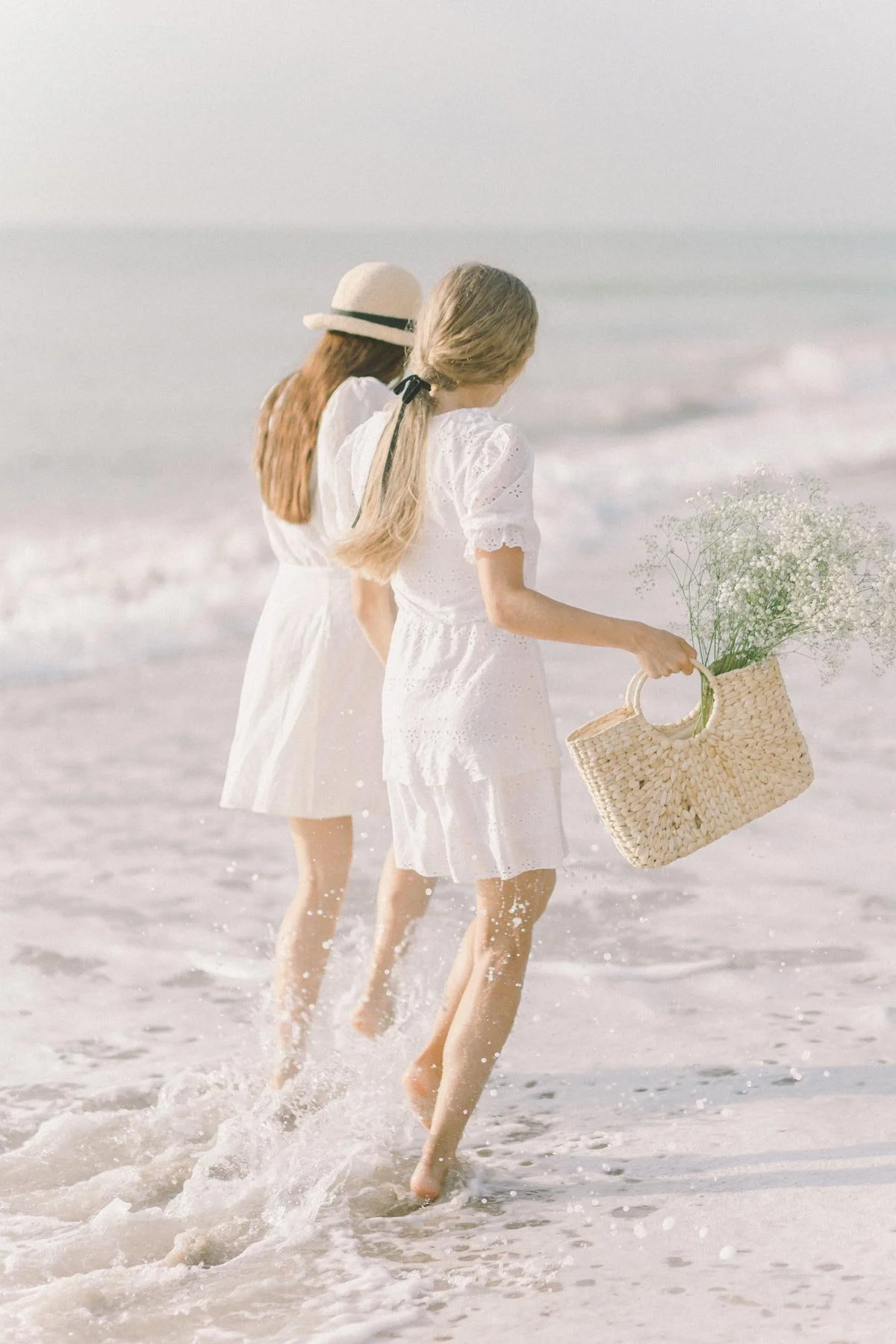 Two women in white dresses walking along the shoreline with waves splashing around their feet, one holding a wicker basket with flowers, at the beach during daytime.