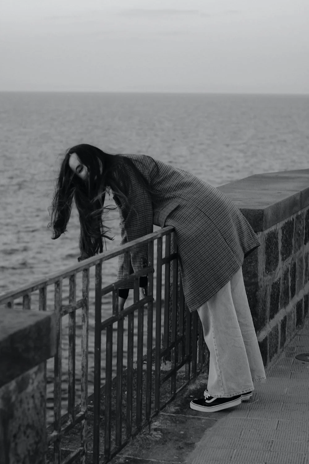 A woman with long hair leaning over a railing near the sea, wearing a checkered coat, light-colored jeans, and sneakers, with wind blowing her hair.