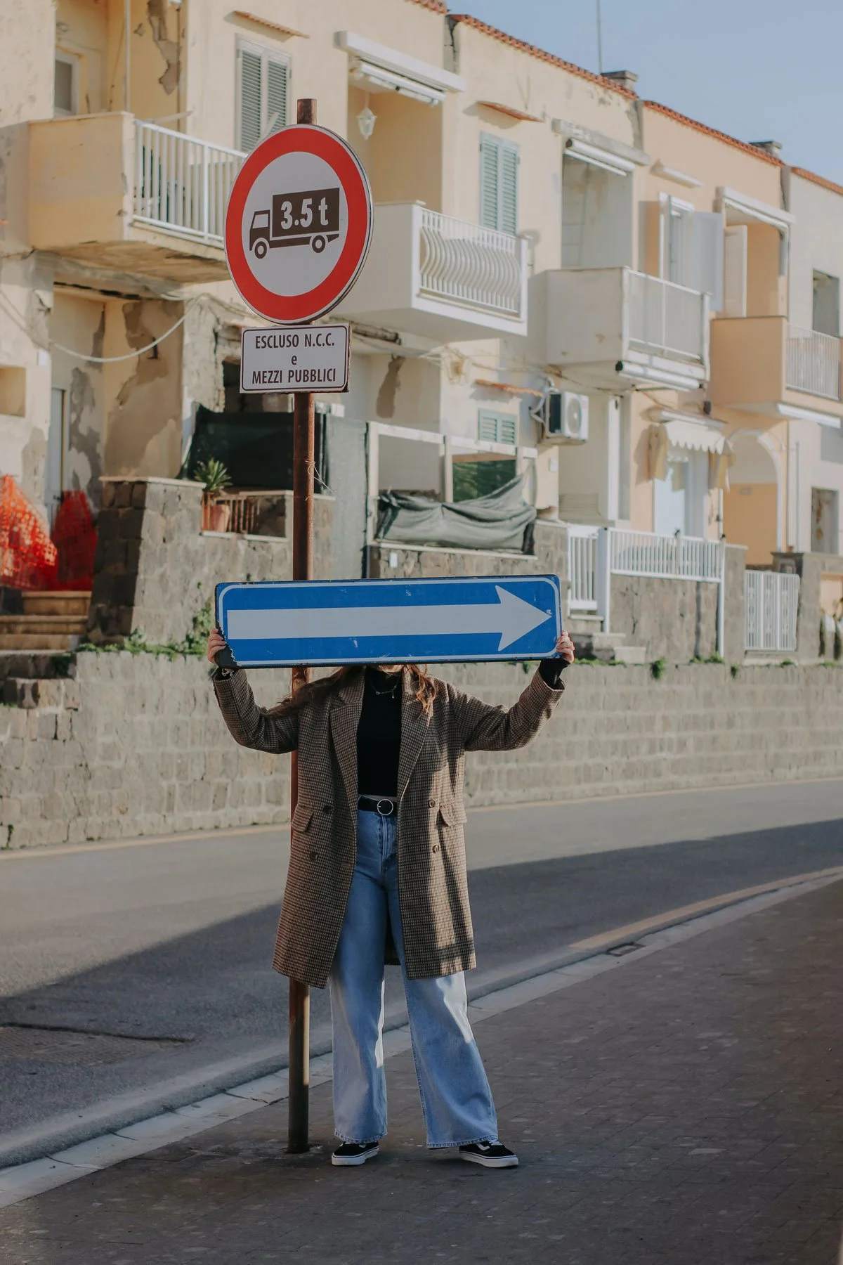 Person holding a blue arrow road sign standing on a sidewalk in front of a street with residential buildings.