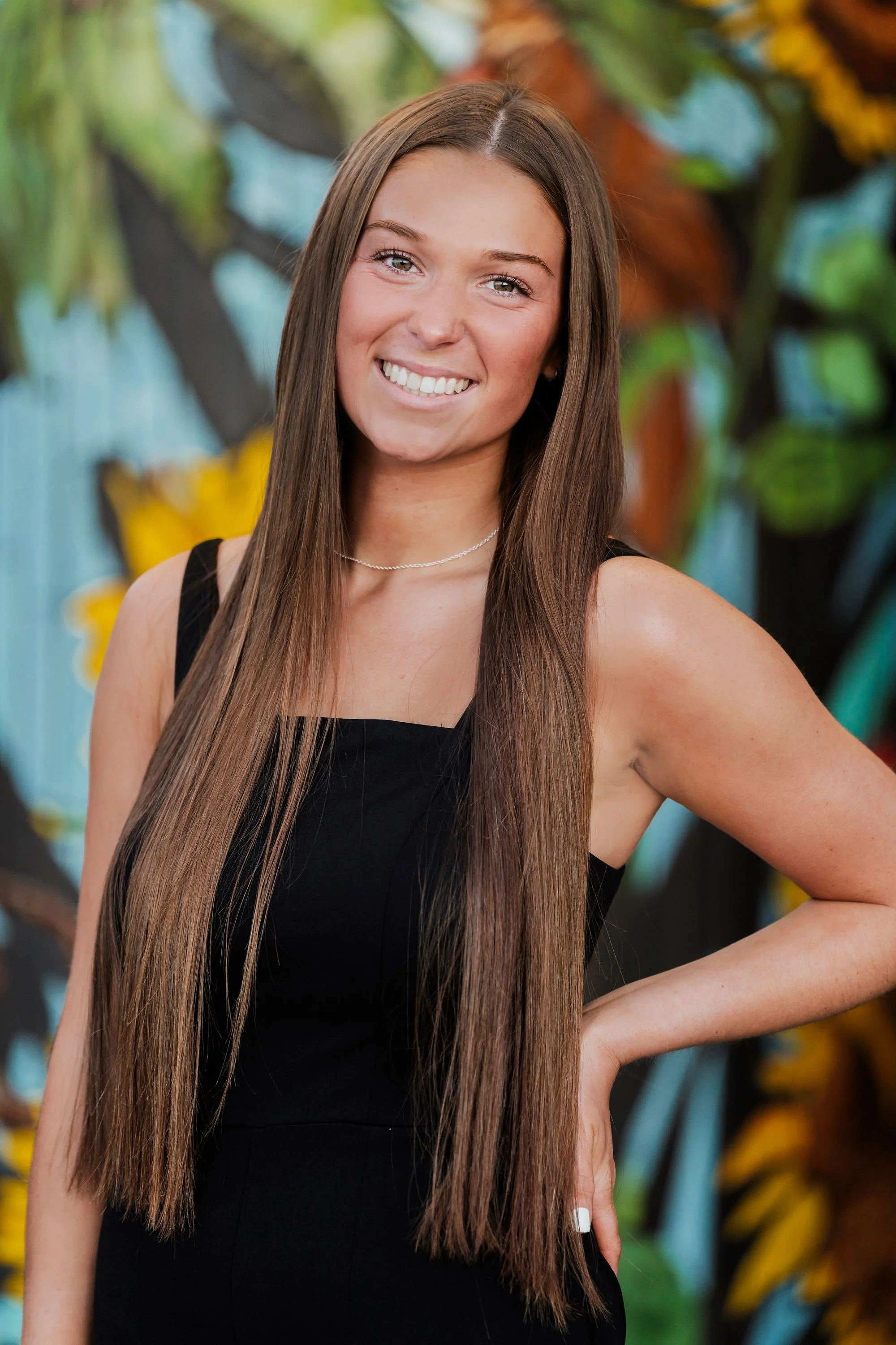 A young woman with long straight brown hair smiling and posing with her hand on her hip in front of a colorful, leafy background.