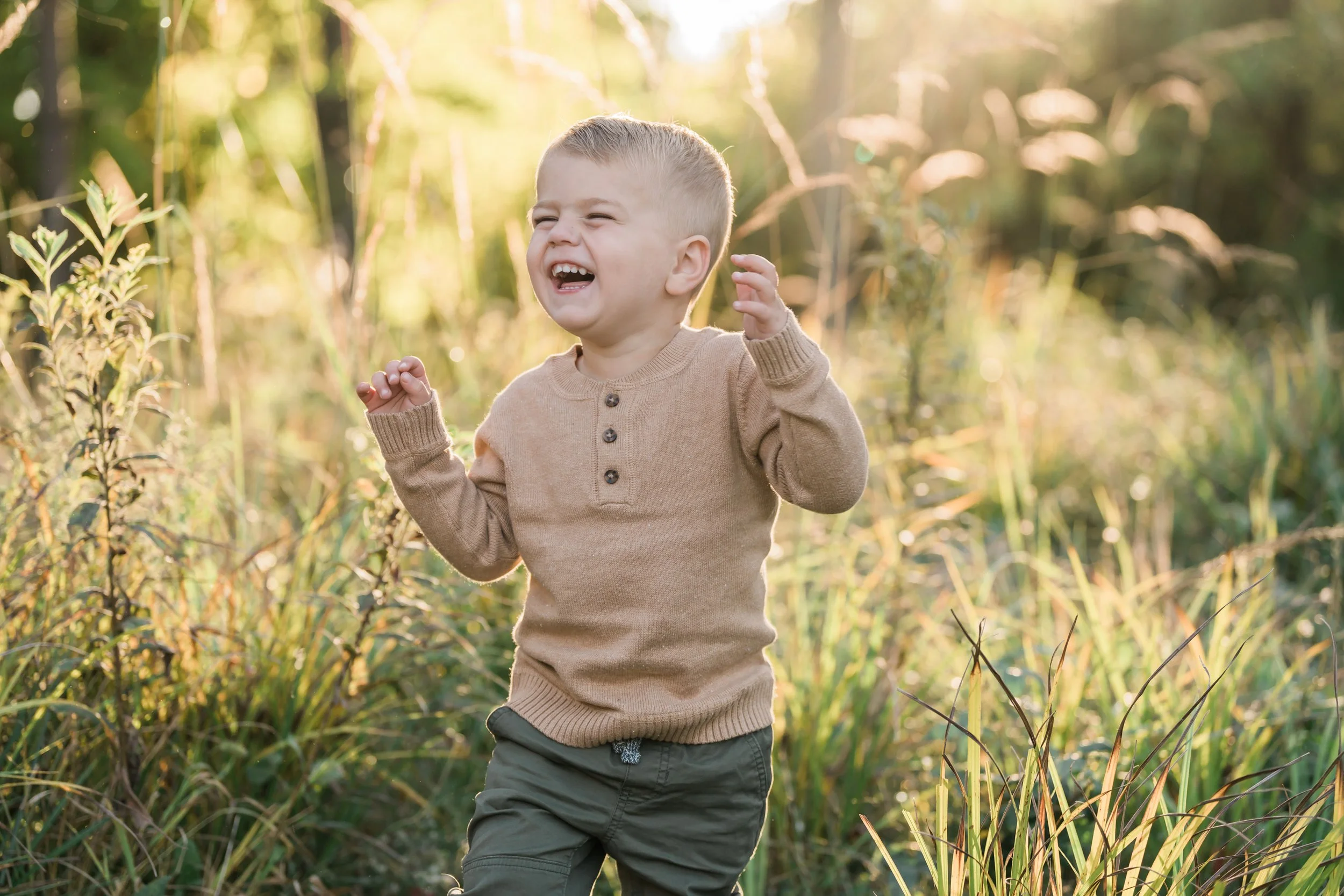 A young boy wearing a beige sweater and green pants is running through a grassy field with tall plants, smiling and laughing with sunlight shining behind him.