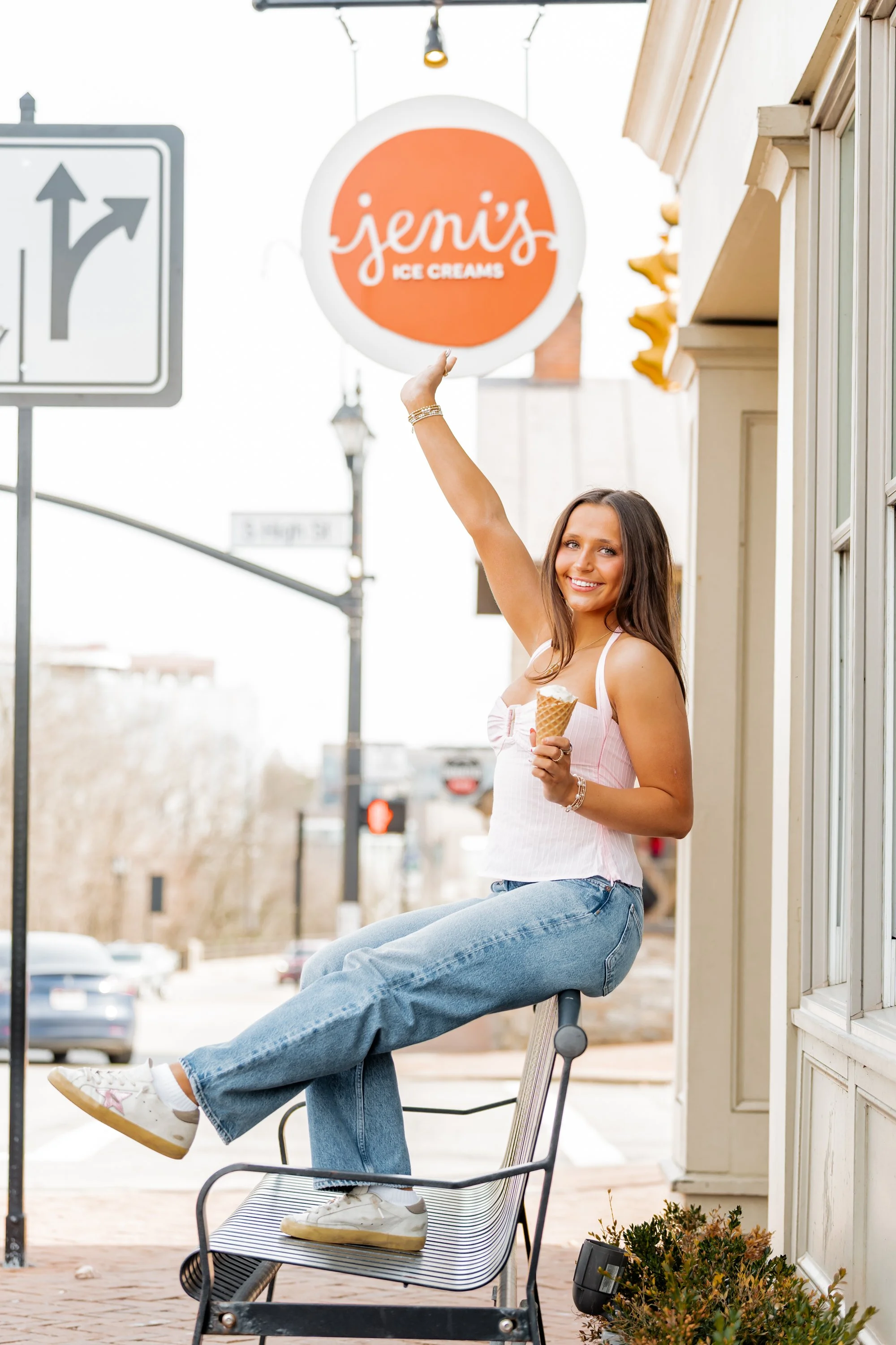 A young woman sitting on a chair outside an ice cream shop, holding an ice cream cone in one hand and reaching up with the other hand towards a round orange sign that reads 'Jeni's Ice Creams,' smiling.