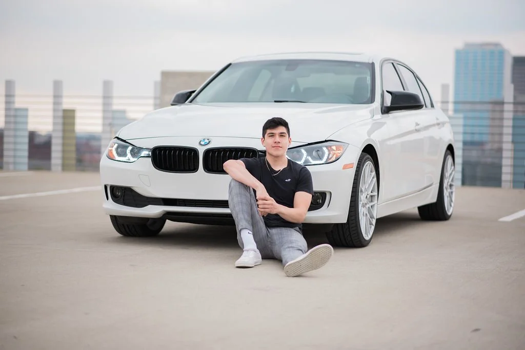 A young man sitting on the ground in front of a white BMW car on a rooftop parking lot with city buildings in the background.